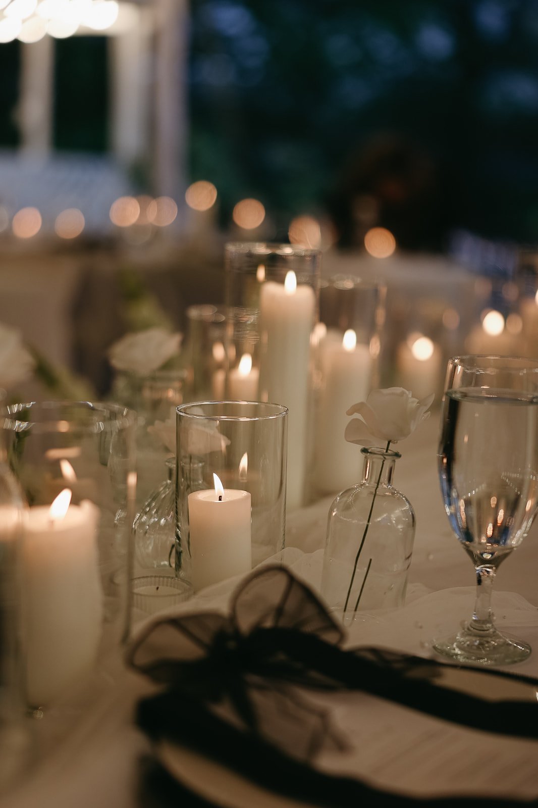Table decorated with lit candles in glass holders, white flowers in glass bottles, and wine glasses, set for an elegant evening event.