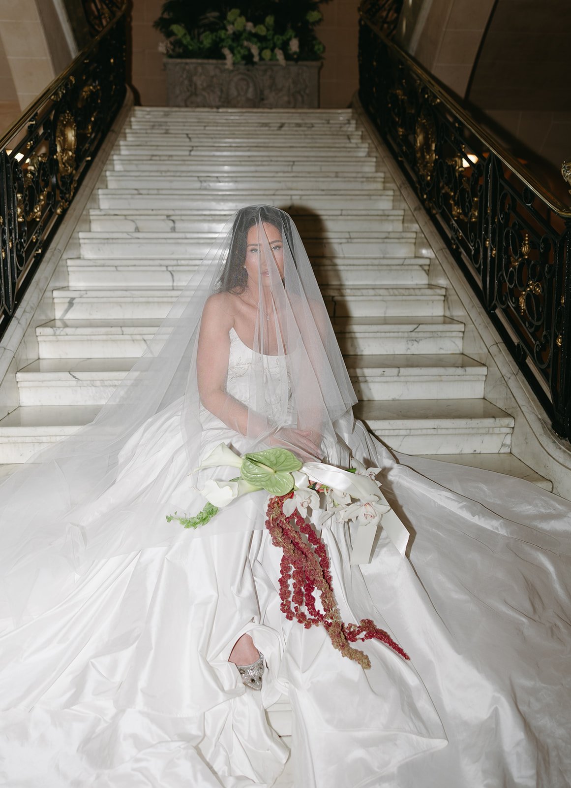 Bride sitting on marble staircase, wearing a white wedding gown, veil, and holding a floral bouquet, with ornate black and gold railing on either side.