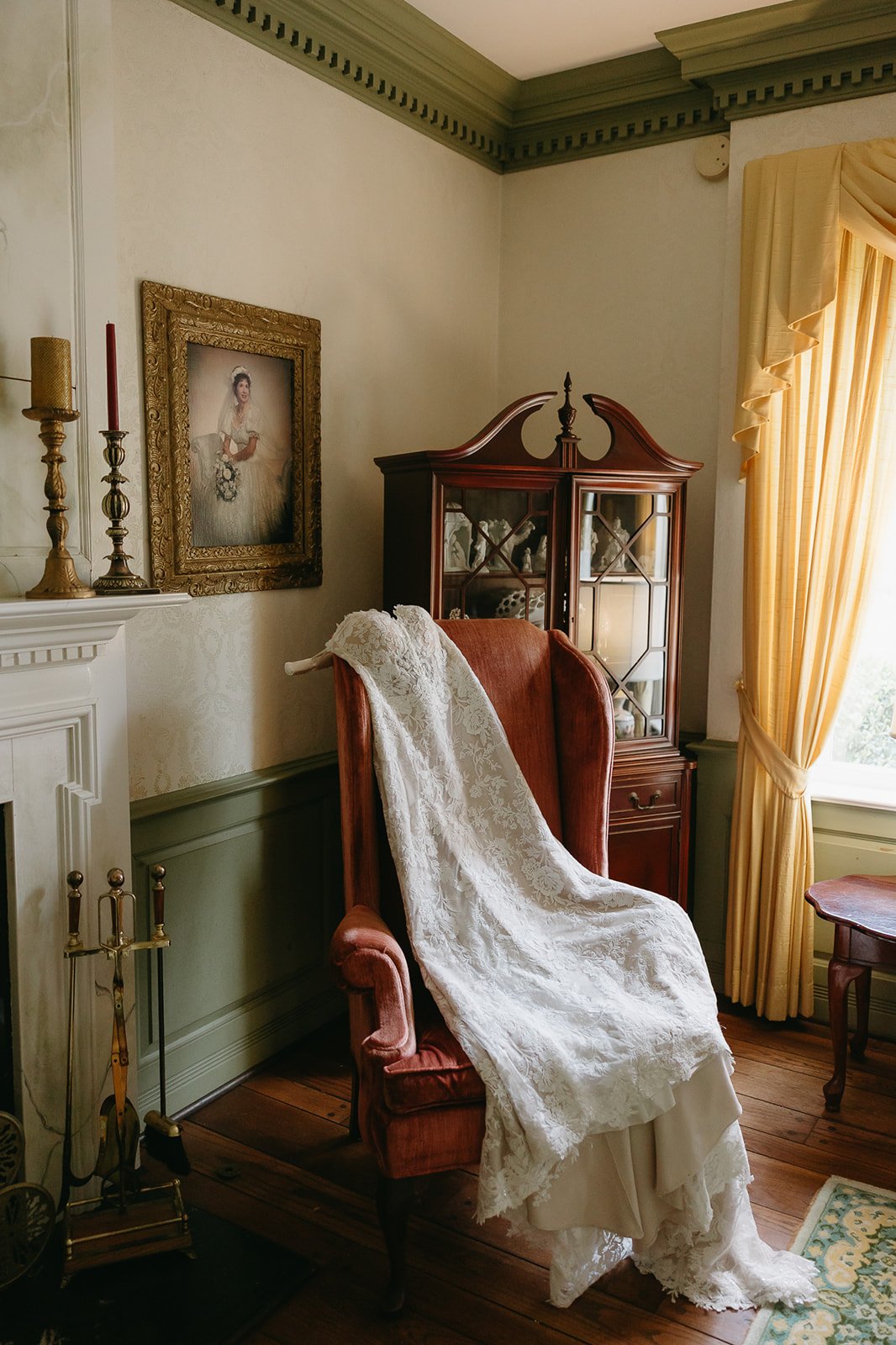 A vintage cozy room with a fireplace, framed painting of a woman, two candlesticks with candles, a wooden display cabinet, an armchair with a white lace wedding dress draped over it, yellow curtains, and hardwood flooring.