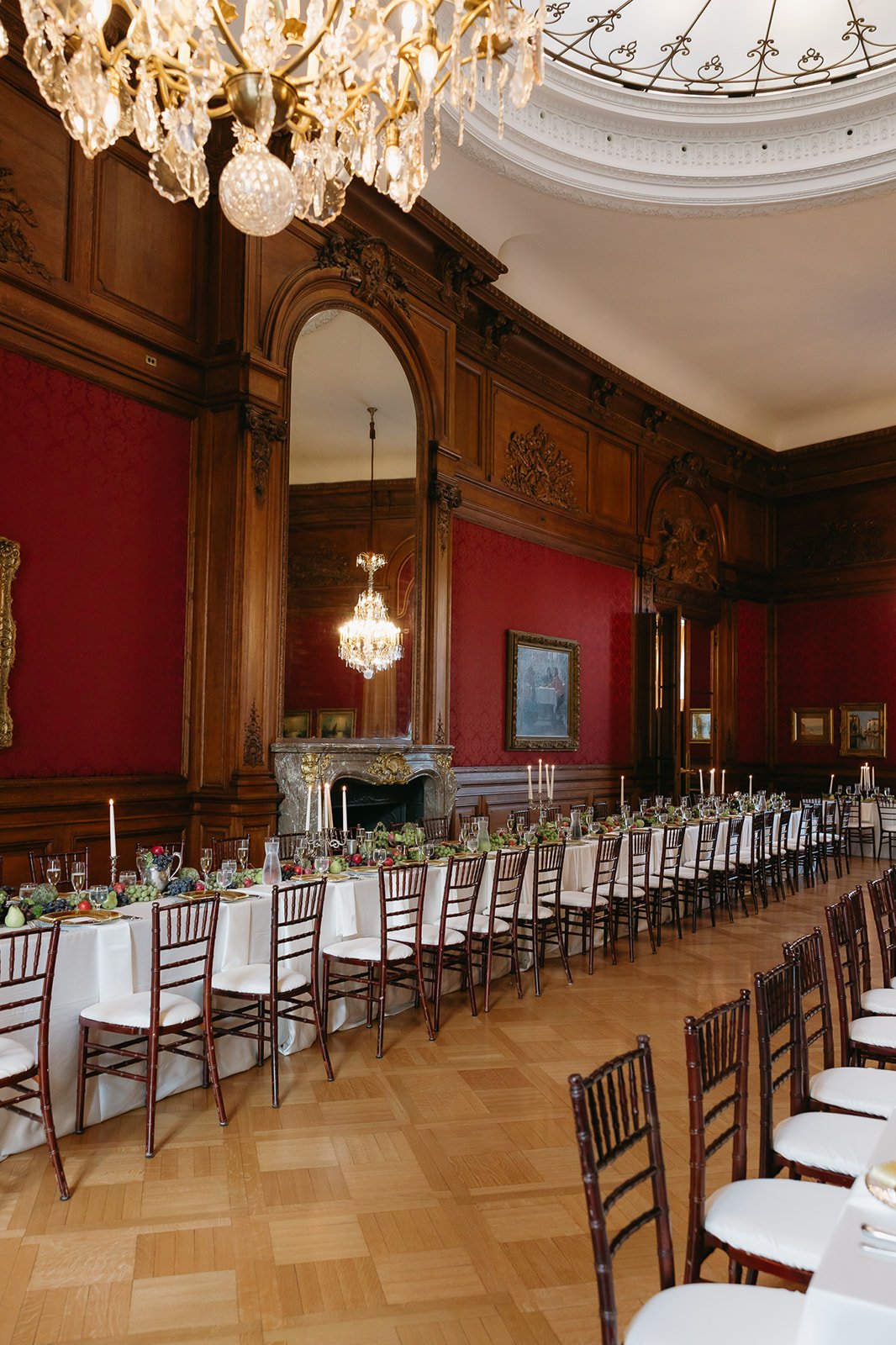 Elegant banquet room with a long table set with white tablecloths, candles, and decorations, surrounded by wooden chairs, ornate wood-paneled walls, marble fireplace, large mirror, chandeliers, and a decorative ceiling.
