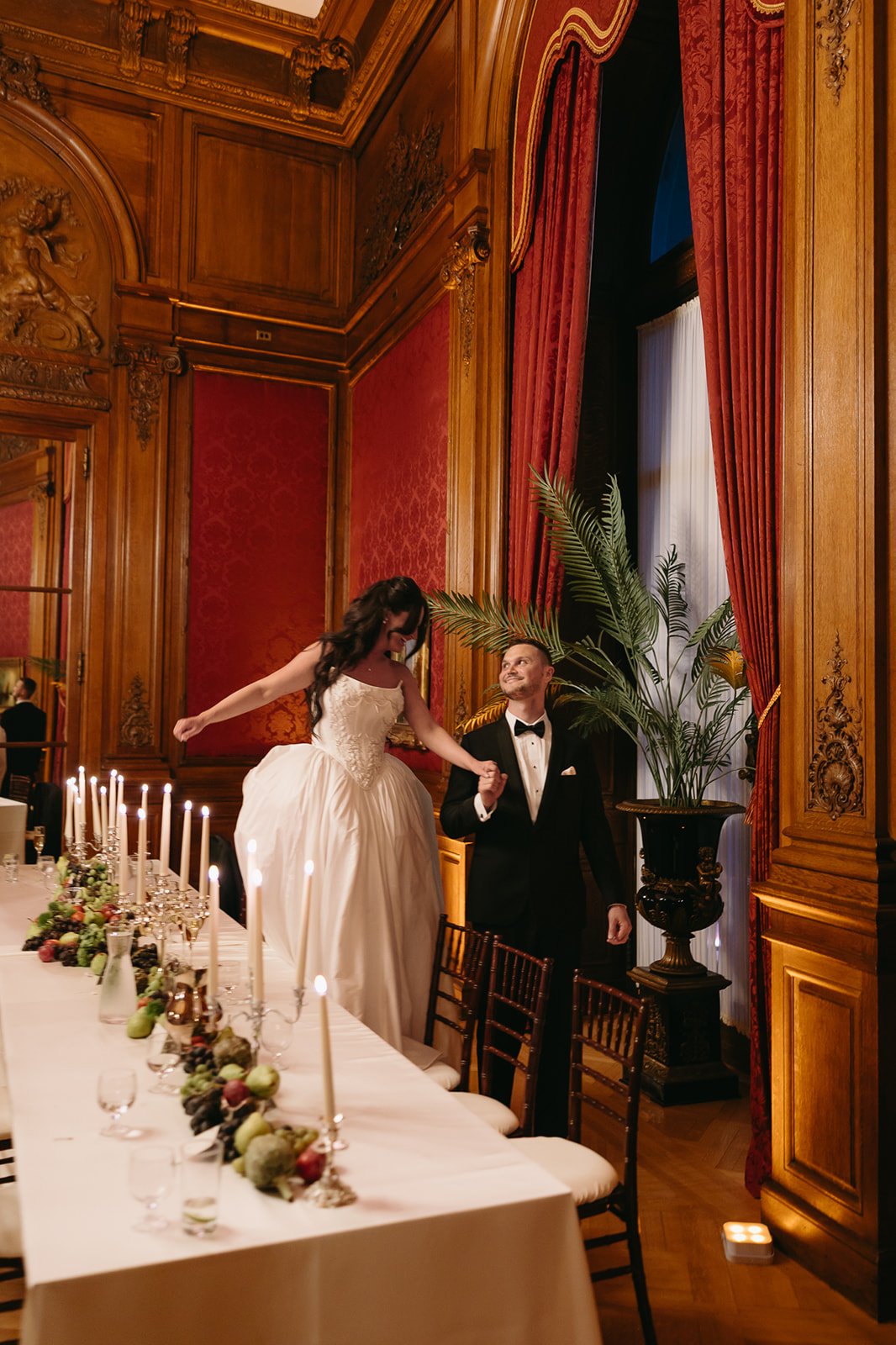 A newlywed couple dancing in a luxurious, ornate room with wood paneling, red wallpaper, and heavy red drapes. The bride is wearing a white wedding gown and is being lifted by the groom in a tuxedo. The room has a long reception table with candlestic