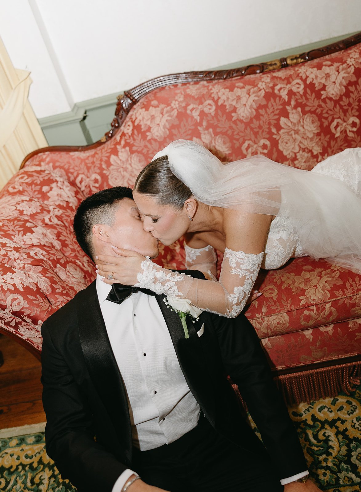 Bride and groom sharing a kiss on a vintage red floral sofa, indoor setting with hardwood flooring.