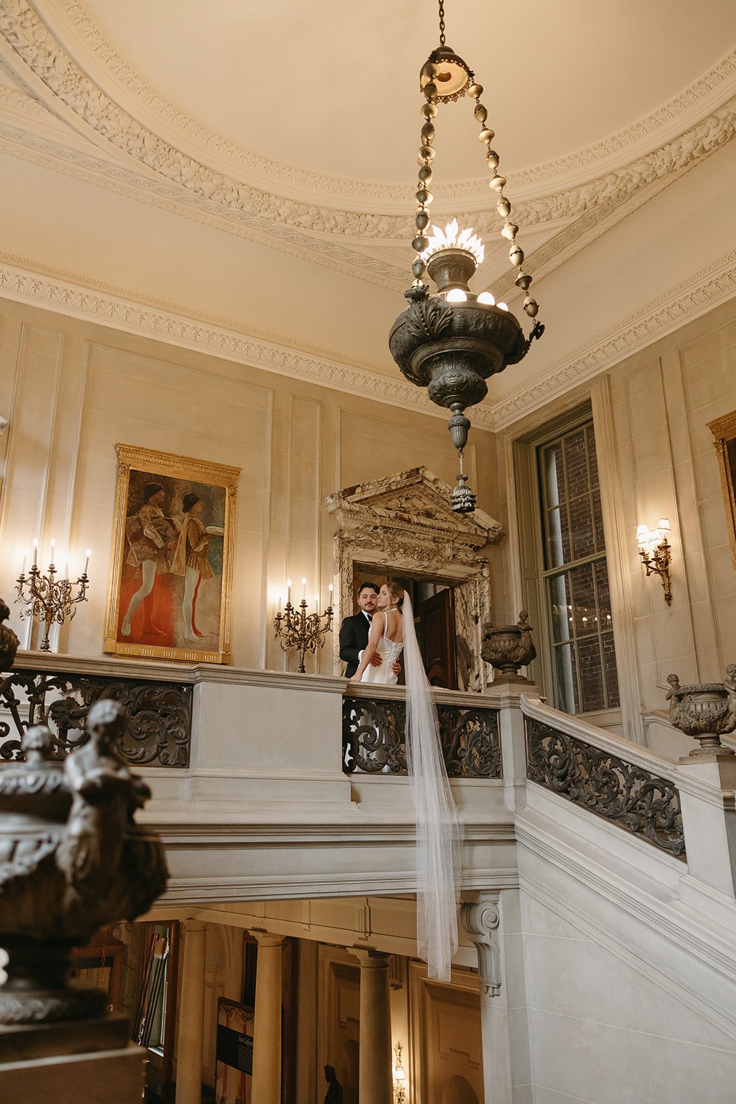 A bride and groom standing on a staircase in an elegant, historic building with ornate details, large windows, and vintage chandeliers.