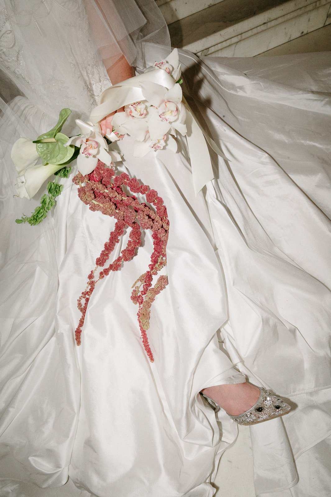 Close-up of a wedding dress with floral embellishments and a bouquet of white flowers, including orchids and calla lilies, resting on the dress. A single silver high-heeled shoe is visible near the bottom.