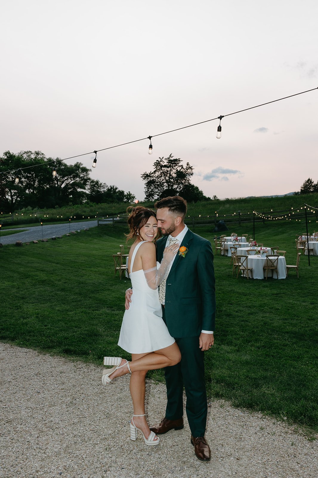 A couple at their outdoor wedding reception, dressed in wedding attire, standing on a gravel pathway with decorated tables in the background and string lights hanging above during dusk.