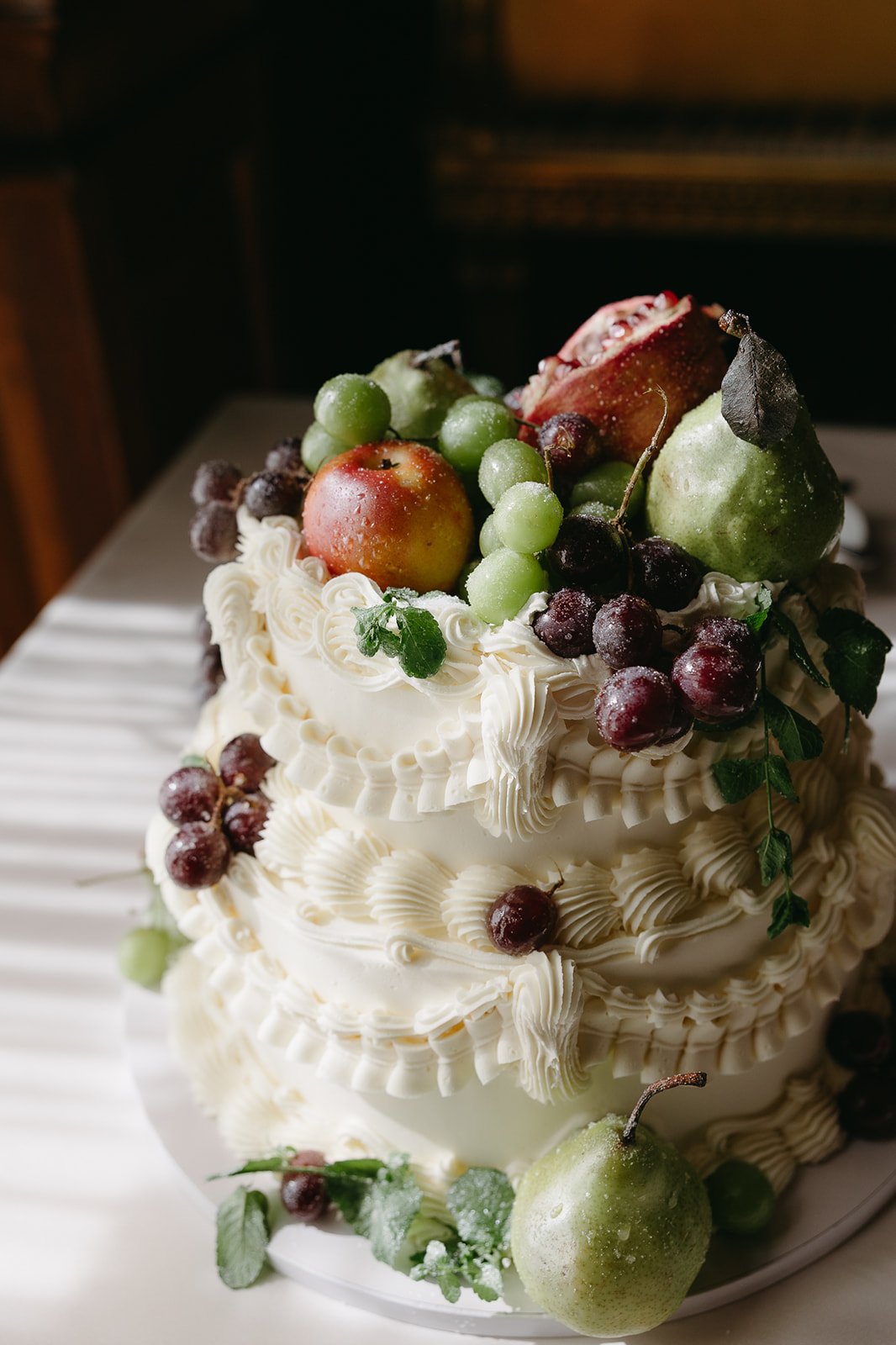 A two-tiered white cream cake decorated with fresh green and red grapes, apples, and pears, with piped cream designs and leaves, set on a white plate.