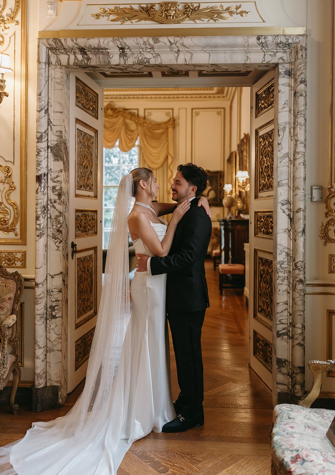 A bride and groom dancing in an elaborately decorated room with gold and marble details, warm lighting, and large windows in the background.