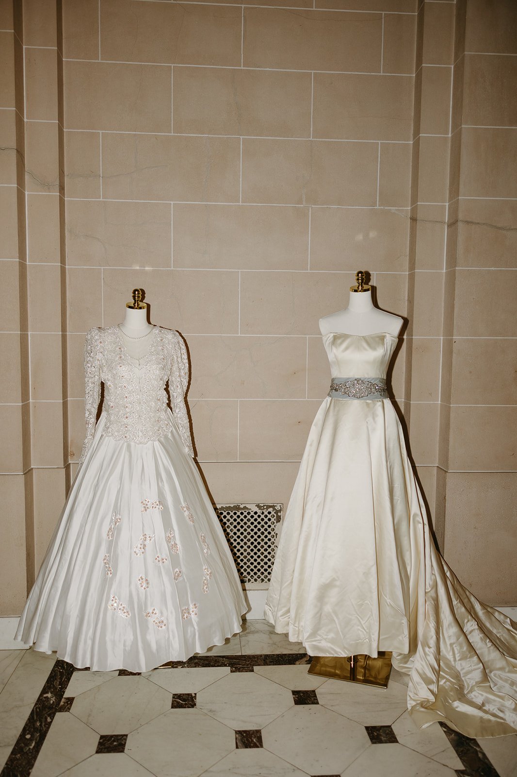 Two wedding dresses on mannequins displayed against a beige tiled wall.