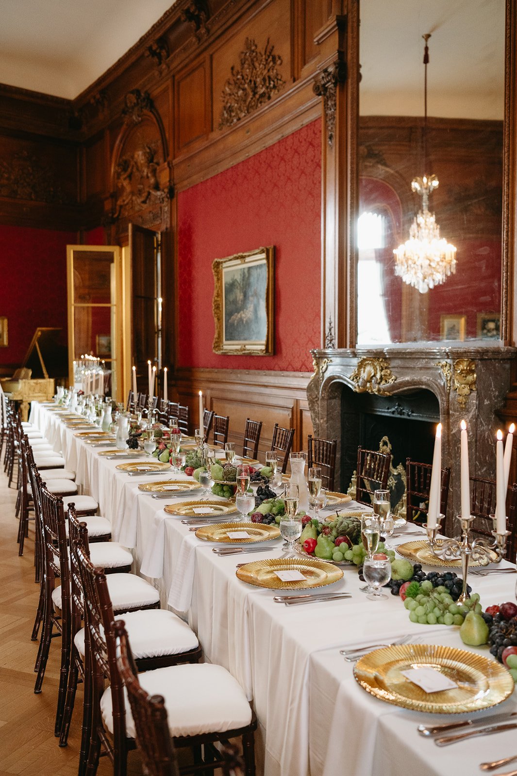 Long dining table set for a formal event with gold chargers, white napkins, glasses, and a centerpiece of grapes, apples, and pears in a luxurious room with red walls, wooden paneling, a fireplace, and a chandelier.