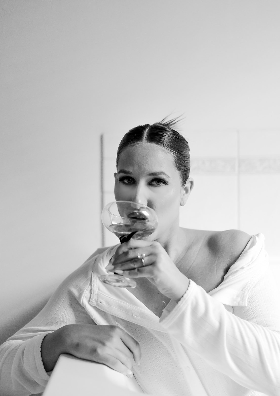 A black-and-white photo of a woman with slicked-back hair, wearing a white shirt, holding a cocktail glass near her lips, looking directly at the camera.