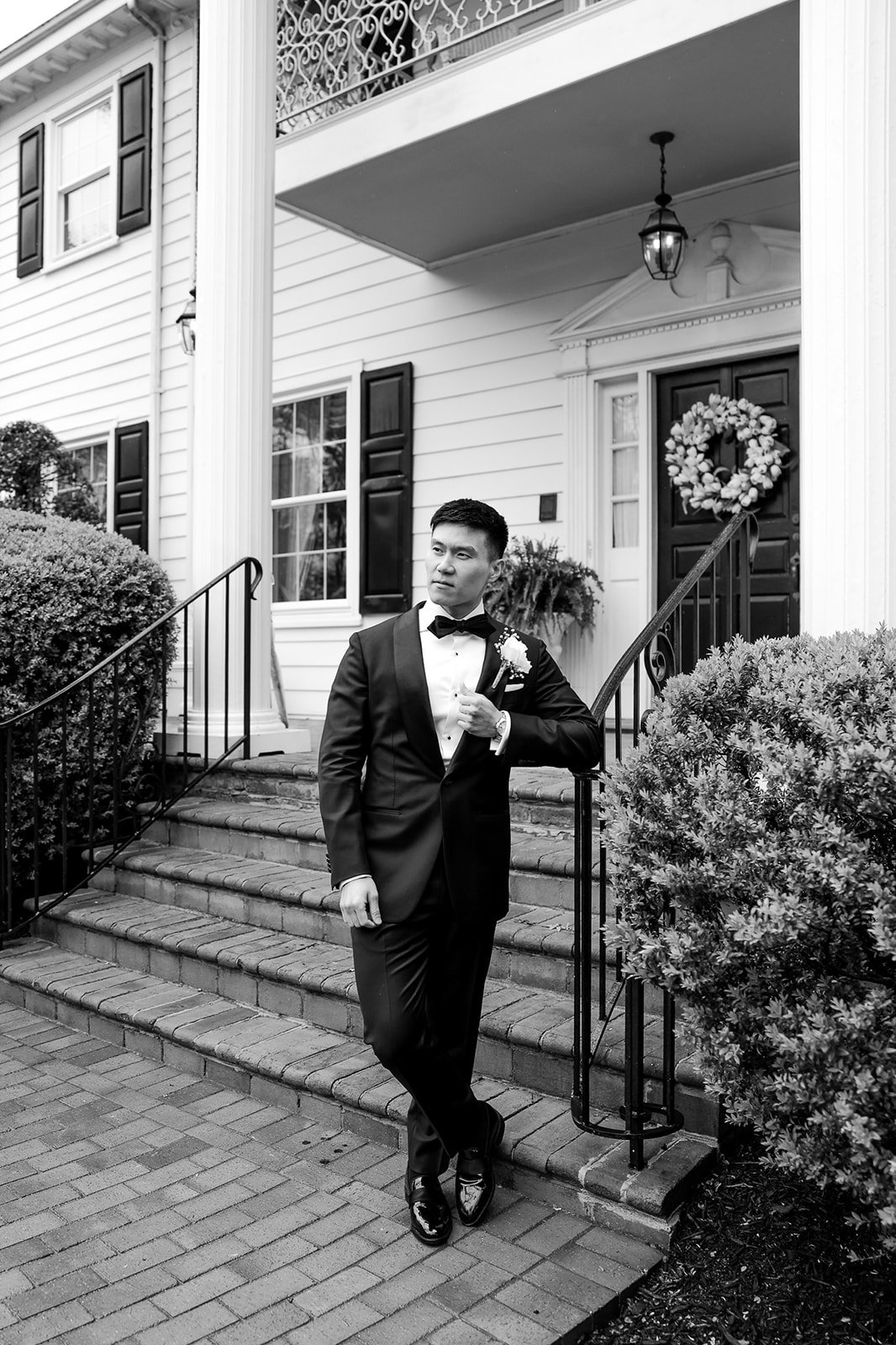 A man in a tuxedo standing on the front steps of a house adorned with a wreath, surrounded by bushes.