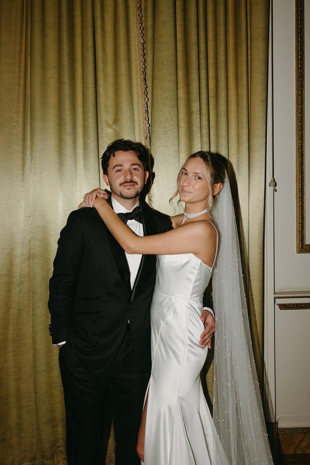 A bride and groom pose together at their wedding indoors with gold curtains in the background. The groom wears a black tuxedo with a bow tie, and the bride wears a white satin wedding gown with a slit and a veil.