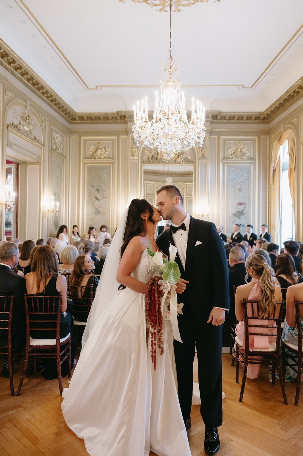 A bride and groom kissing at their wedding ceremony in an elegant, decorated ballroom filled with seated guests.