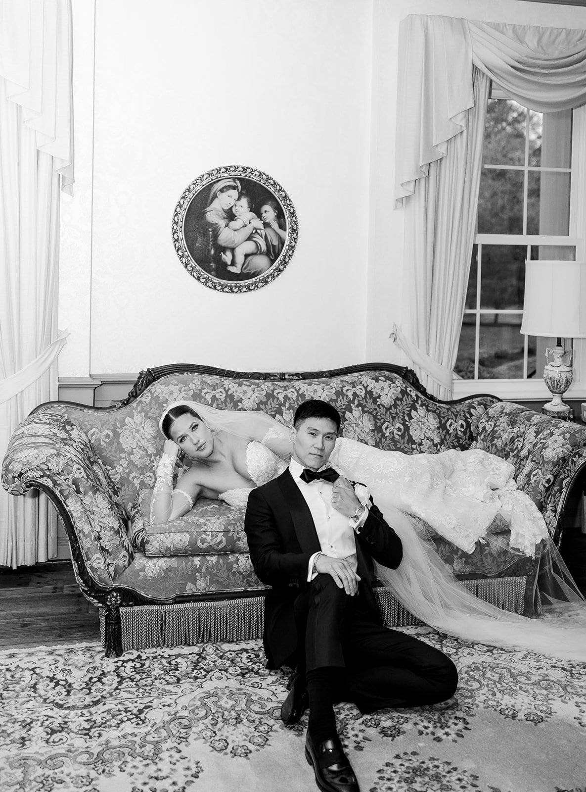 Black and white photo of a bride in a wedding dress reclining on a floral sofa and a groom in a tuxedo kneeling in front of her with one knee on the rug, dressed in a tuxedo with a bow tie, inside a room with large windows, curtains, and a picture on