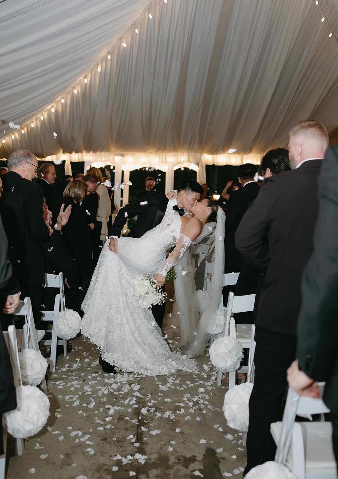 Bride and groom sharing a kiss at their wedding reception under a white tent, with guests surrounding them, white chairs decorated with flowers, and flower petals on the floor.