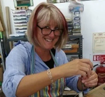 A smiling woman with glasses and blonde hair in a colorful apron is working with her hands in a cluttered workspace.