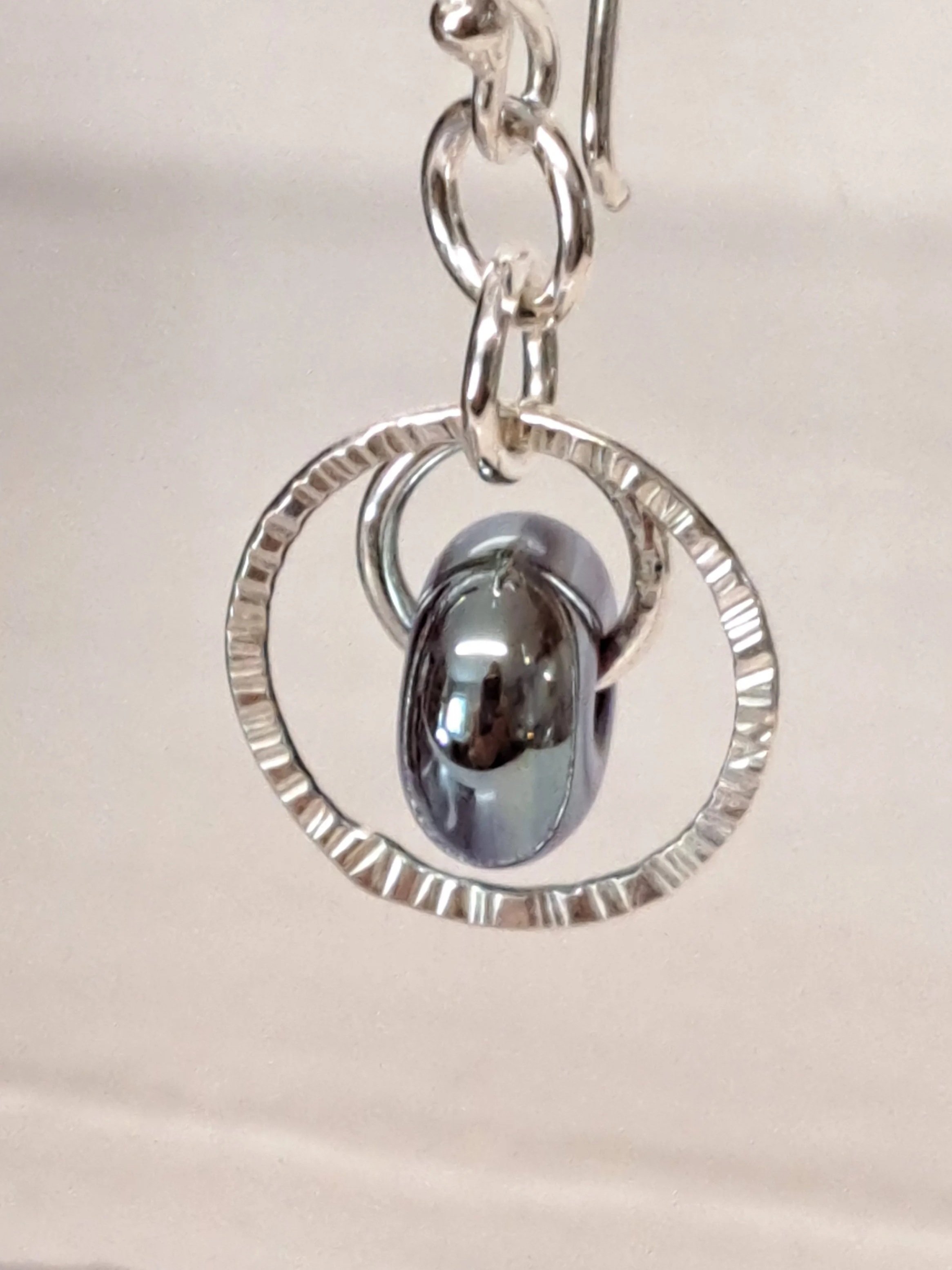 Close-up of silver jewelry consisting of a chain, a circular ring with textured edges, and a polished oval bead or charm hanging in the center.