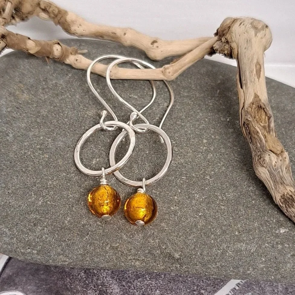 A pair of silver hoop earrings with amber-colored glass beads hanging from them, placed on a flat surface next to a piece of driftwood.
