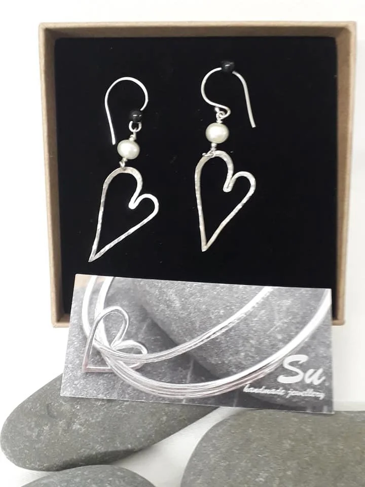 A pair of earrings with heart-shaped metal frames and pearl beads displayed in a jewelry box, accompanied by a necklace and a promotional card with a stone and metal jewelry design.