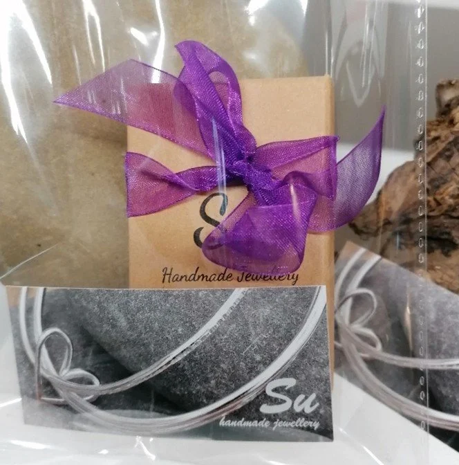 A handmade jewelry card with a purple ribbon tied in a bow, inside a clear plastic bag. The card is beige with black text that reads 'Handmade Jewellery', and part of a black-and-white photo of jewelry is visible.