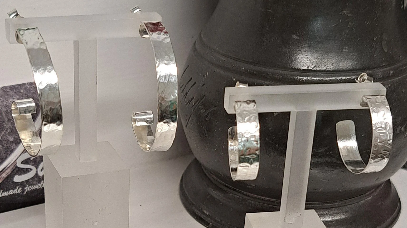 Two pairs of silver bangles displayed on white jewelry stands, set against a large black round object, possibly a decorative urn or vase.