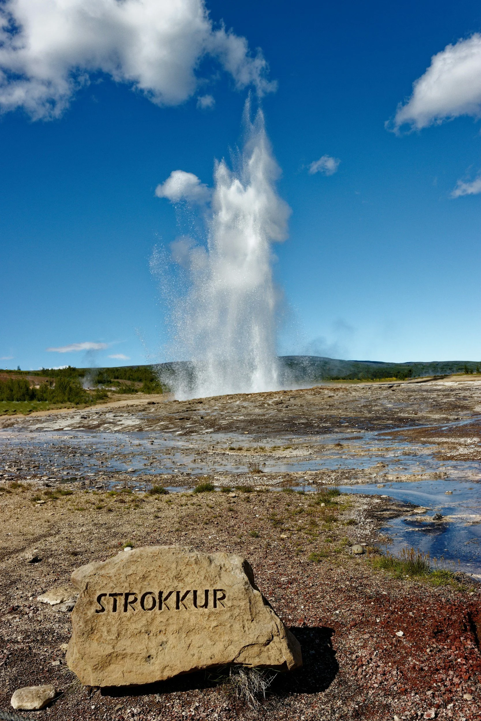 Geyser erupting in a geothermal area with a stone in the foreground inscribed with "Strokkur" and a blue sky with scattered clouds.