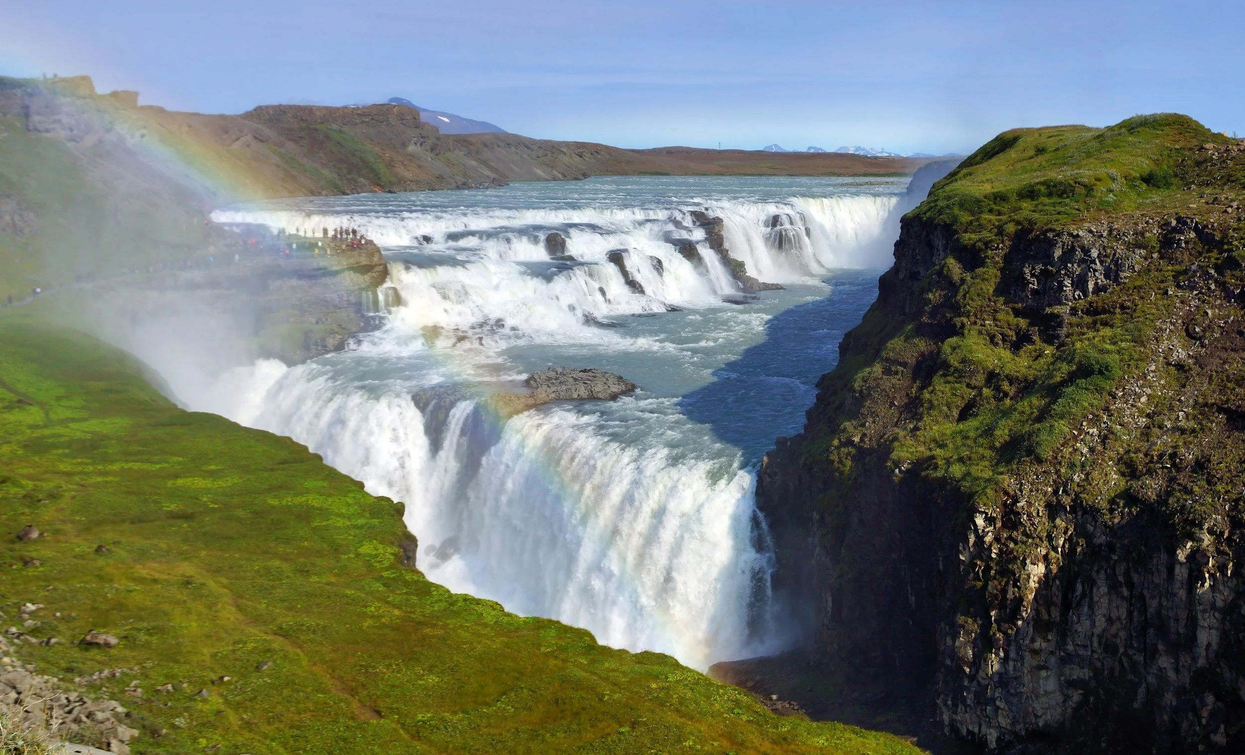 Waterfalls at Gullfoss in Iceland with a rainbow, green grass, and cliffs, under a clear blue sky.