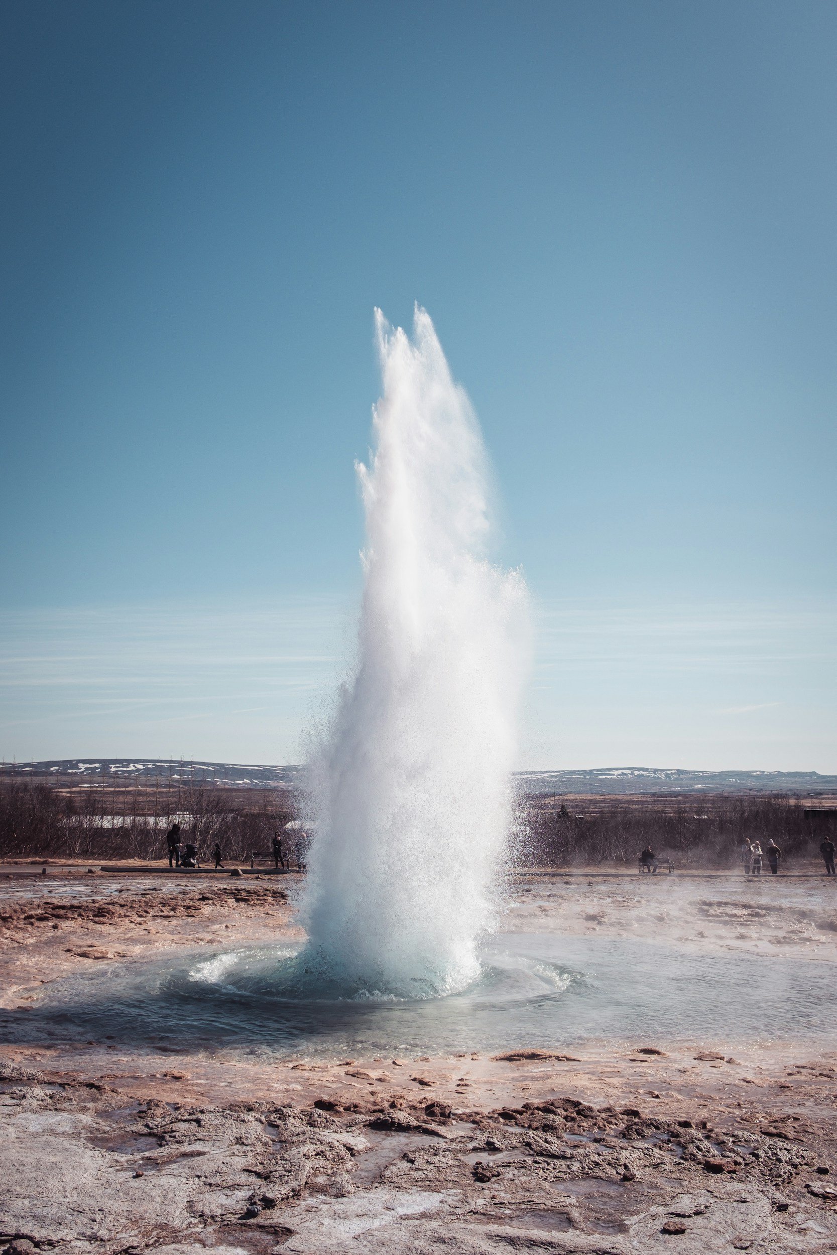 A geyser erupting in a geothermal area with a tall jet of water shooting into the air, surrounded by rocky terrain and people in the background.