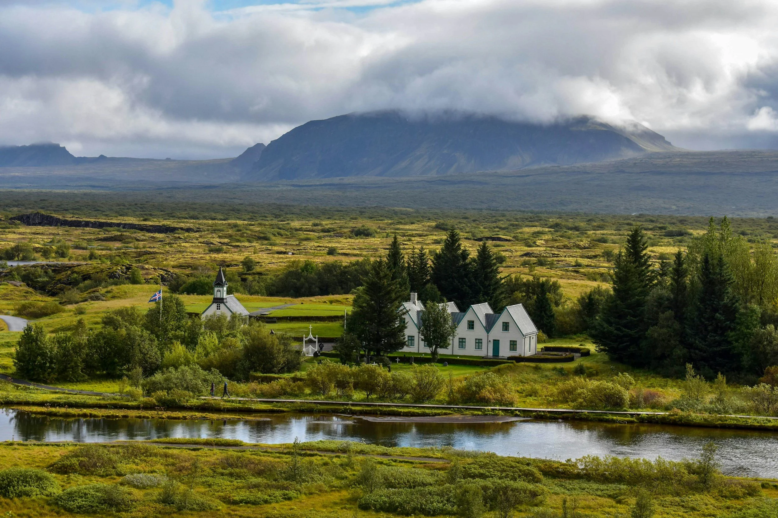A small white church and cottages surrounded by green trees and shrubs, with a pond in the foreground and mountains with cloudy skies in the background.