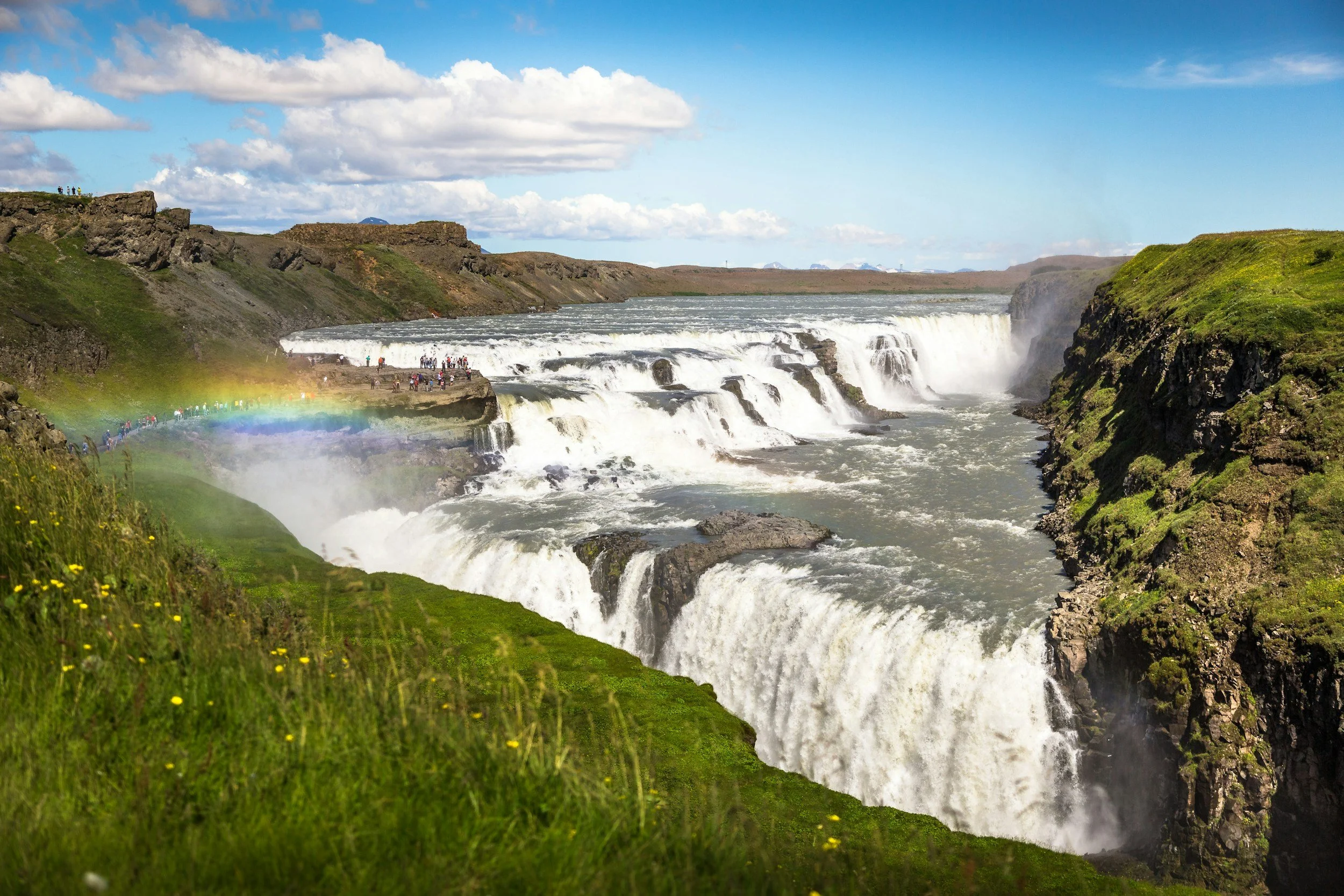 A wide view of Gullfoss waterfall in Iceland with tiers of cascading water, green grassy banks, a rainbow in the mist, and a partly cloudy sky.