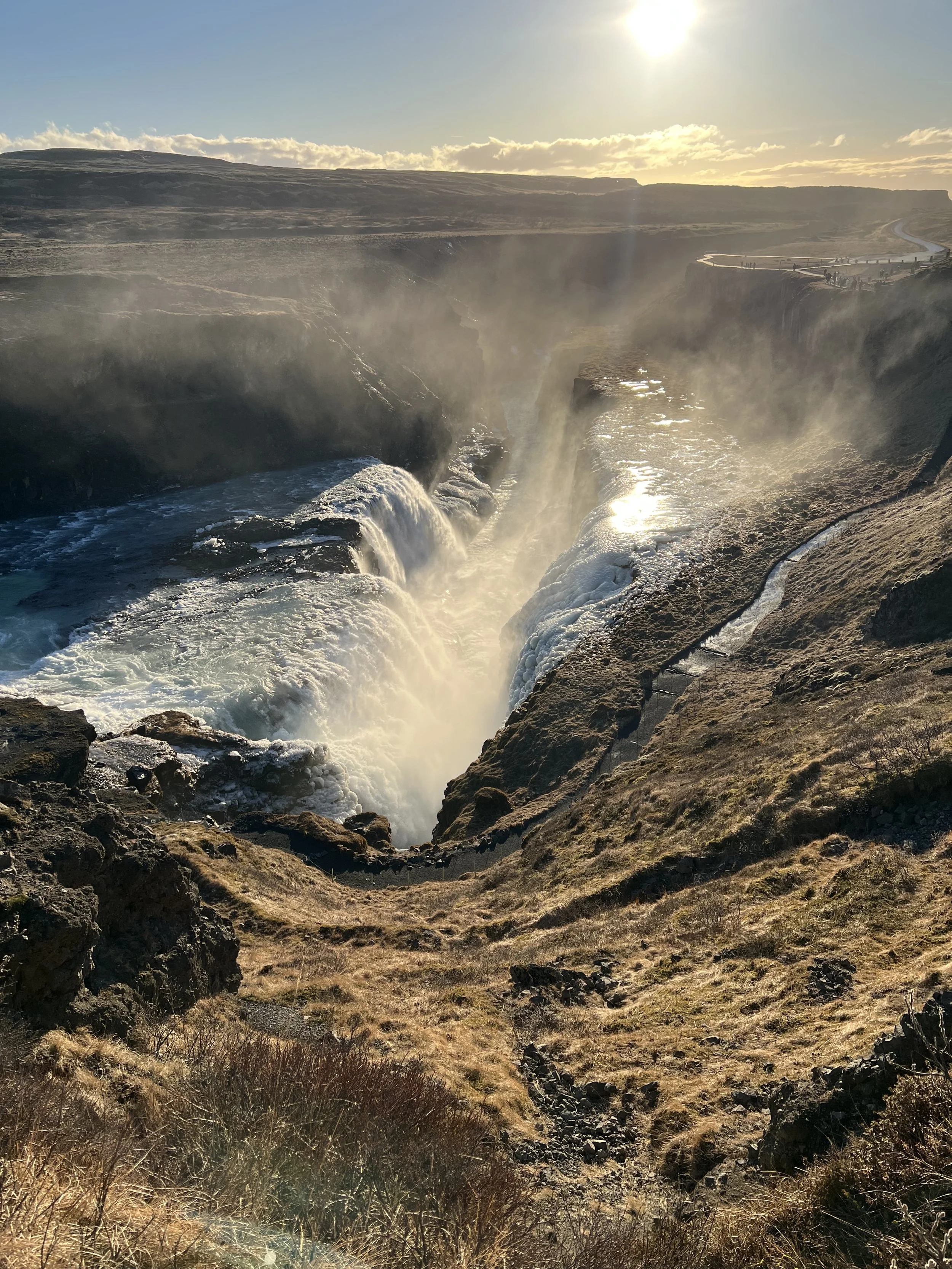 A scenic view of the waterfalls at Gullfoss in Iceland during sunset, with mist rising from the cascading water and a rugged landscape surrounding the falls.