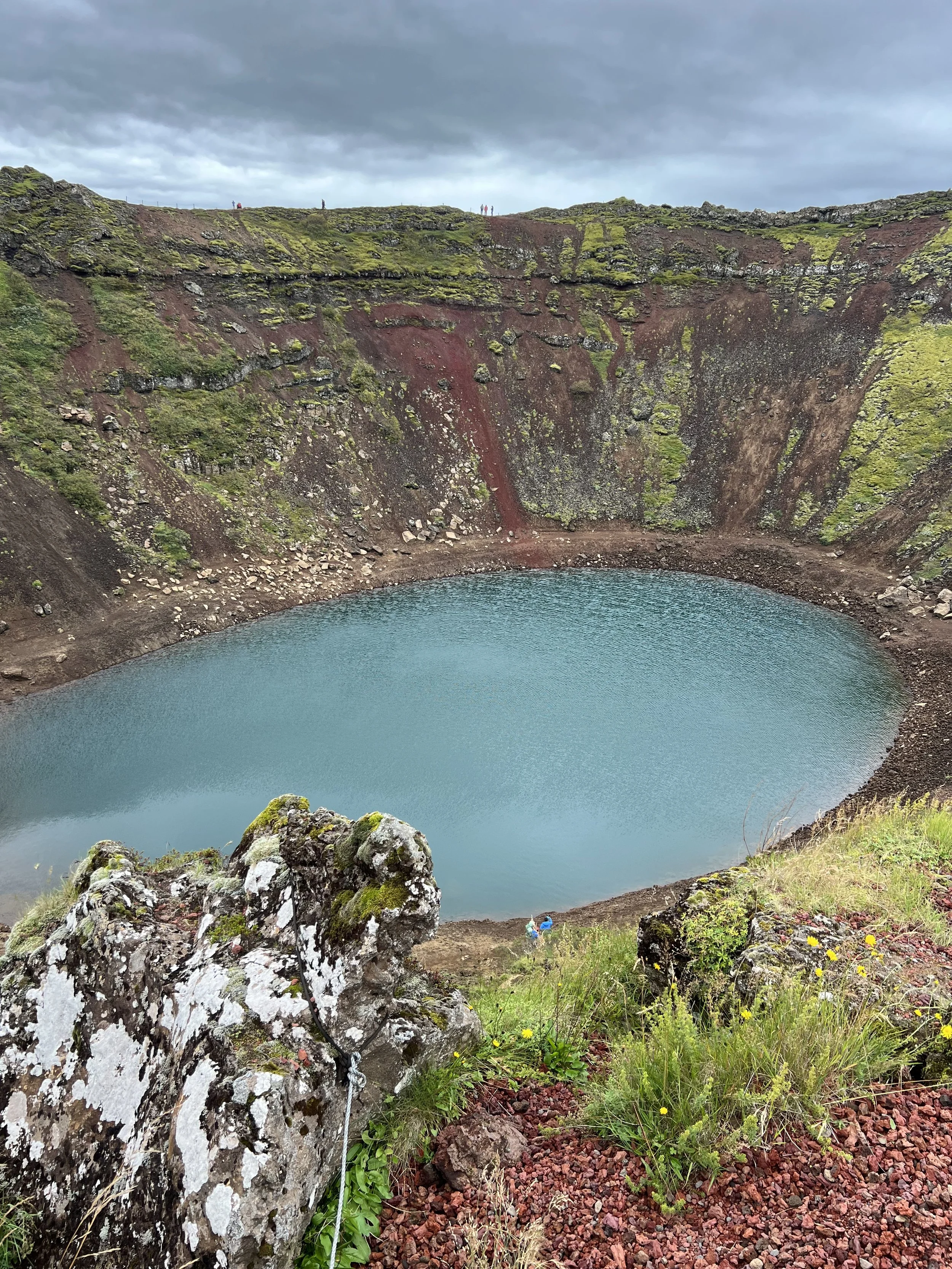 A volcanic crater lake with a steep, moss-covered rim and gray, cloudy sky.