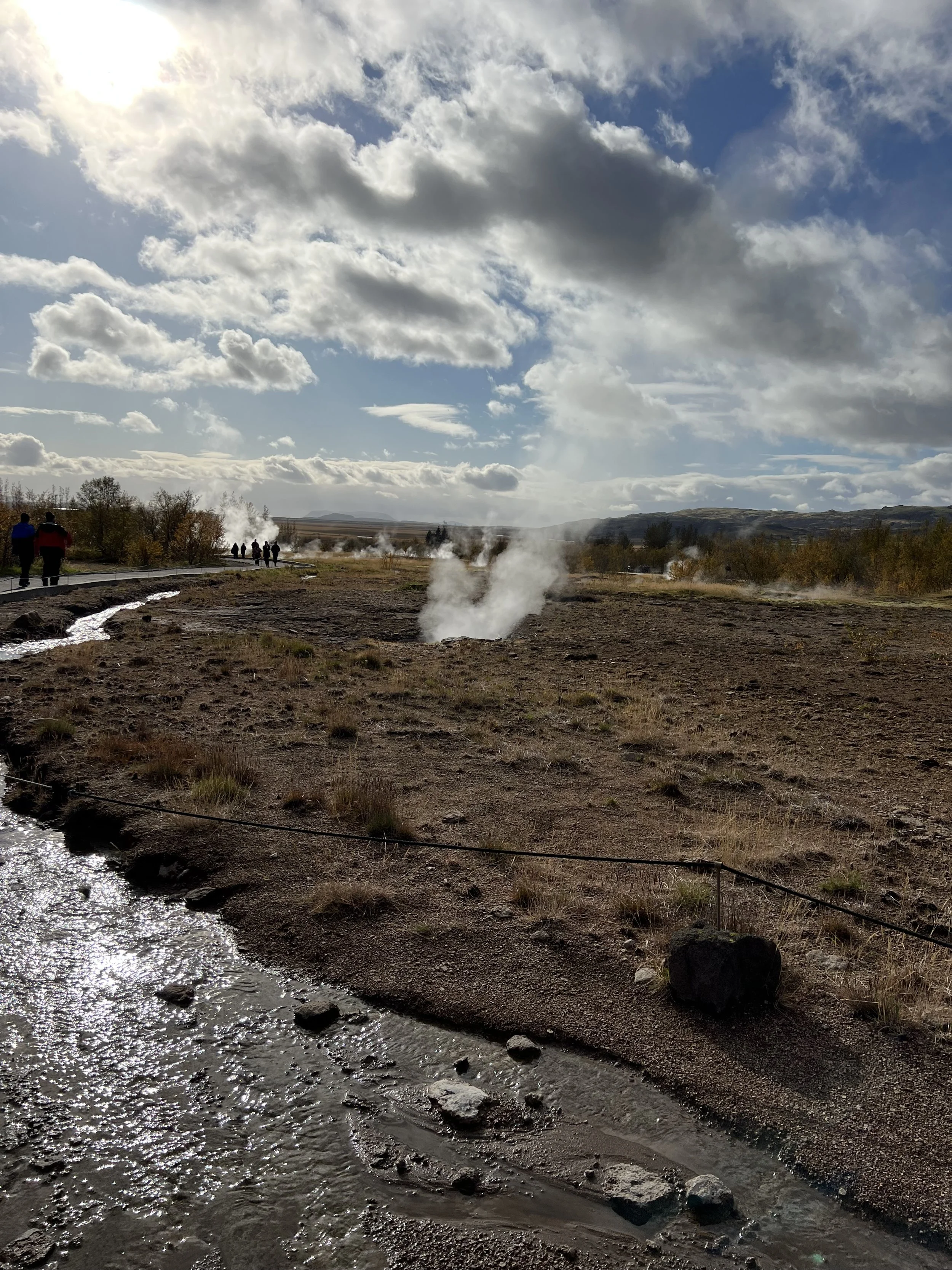 People walking along a path beside steaming geothermal vents and a river under a partly cloudy sky.