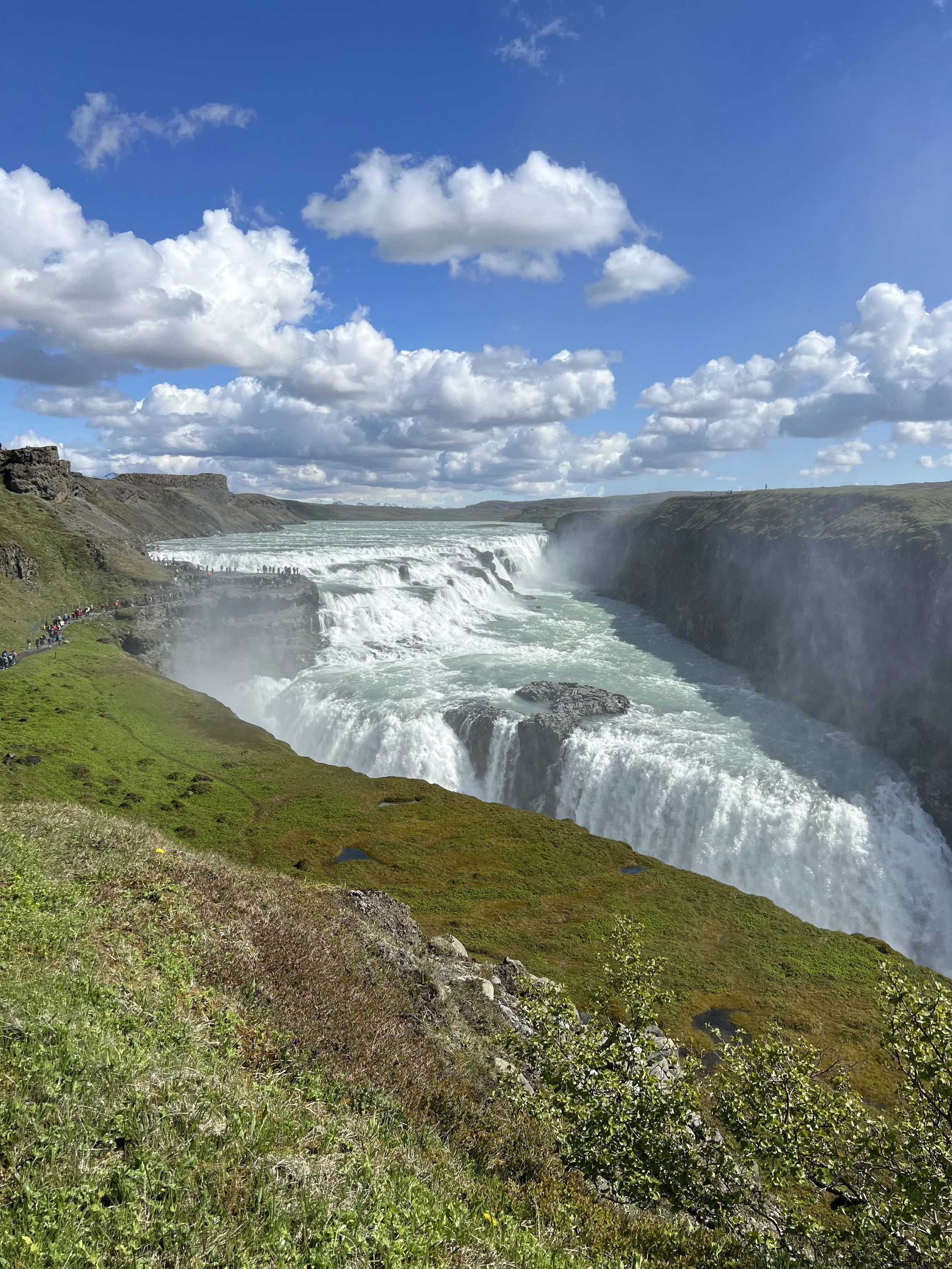 A panoramic view of a Gullfoss with multiple cascades, surrounded by green hills and a partly cloudy blue sky.