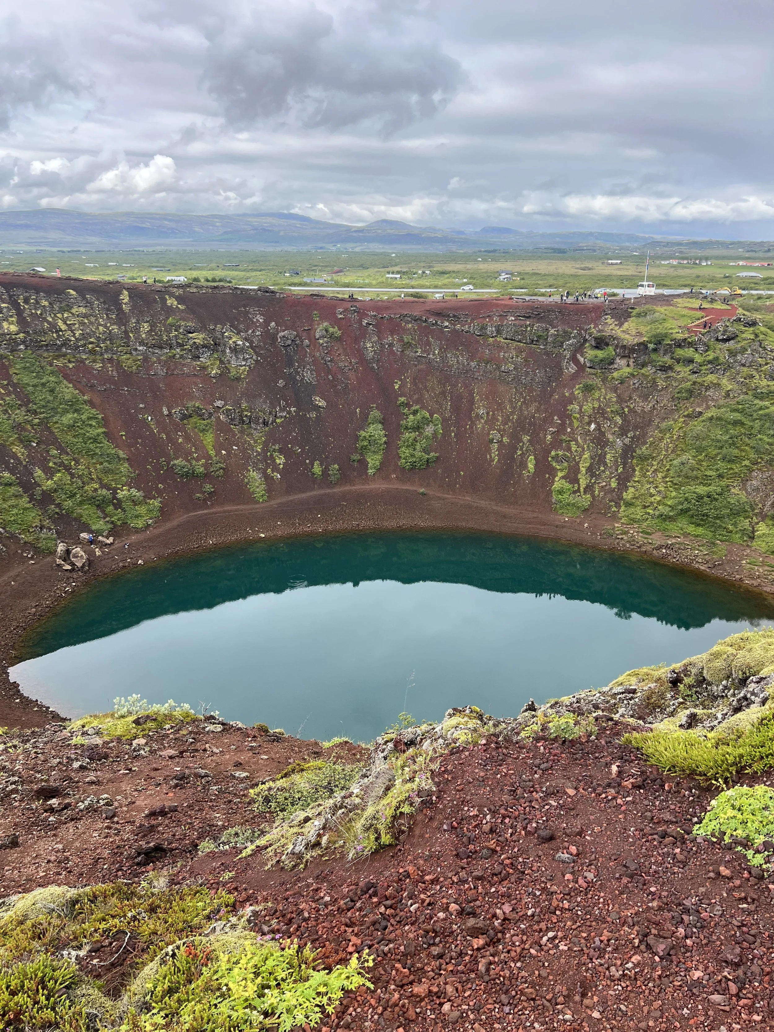 The crater Kerið wiith a teal-colored lake at the bottom, surrounded by red volcanic rocks and patches of green moss and plants. Overcast sky with clouds and distant landscape in the background.