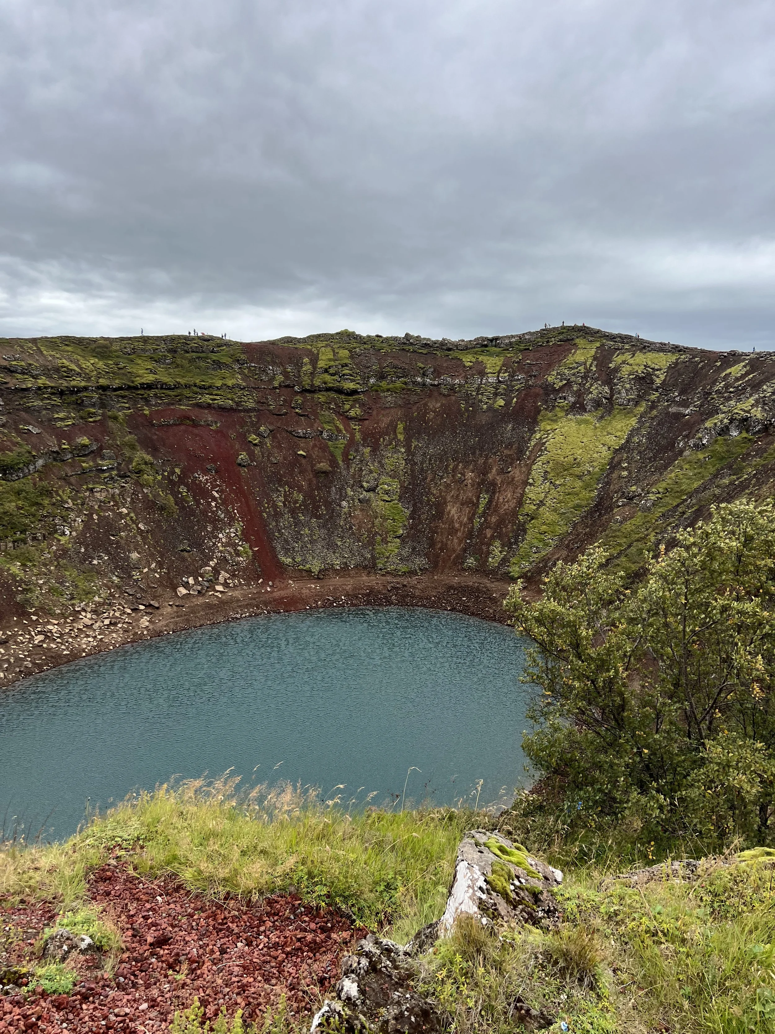 A crater lake surrounded by moss-covered rocky cliffs with green grass and vegetation in the foreground, under a cloudy sky.