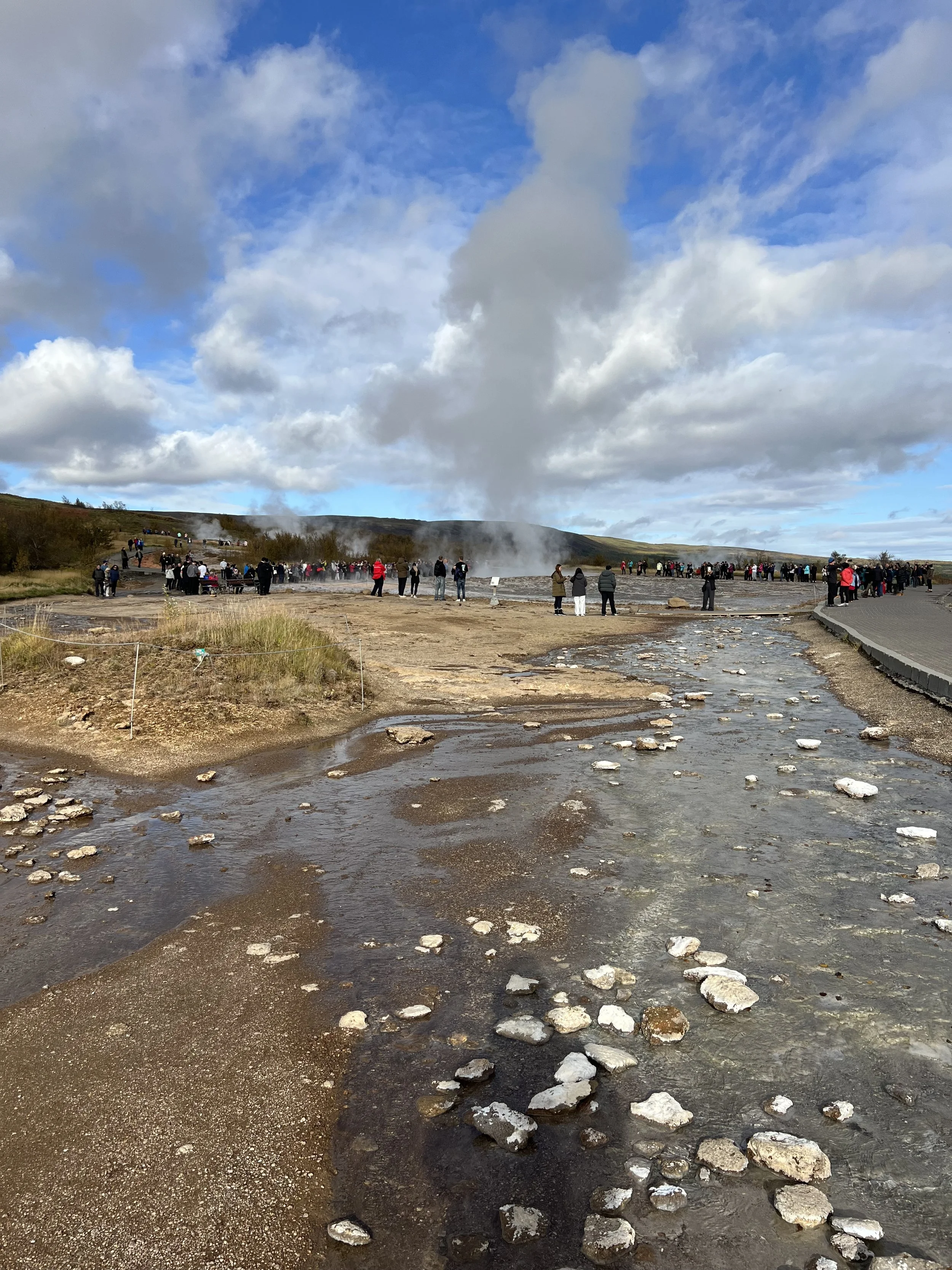 A geothermal area with a steaming hot spring or geyser in the background, surrounded by rocks and a group of tourists walking and observing the scene under a partly cloudy sky.