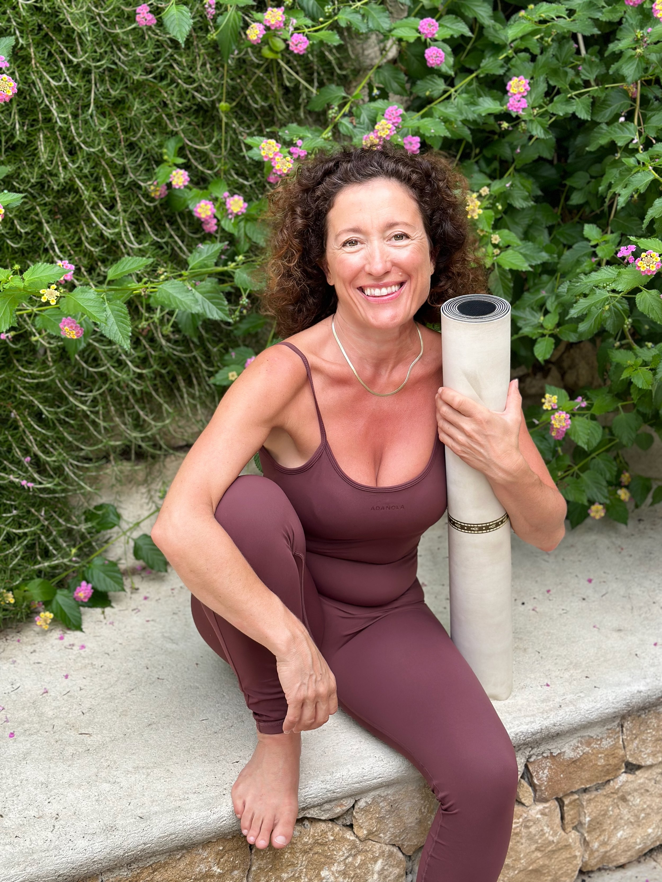 Pilates teacher Bessie Refalo smiling and holding her mat at a Greek retreat, inviting new clients to connect.