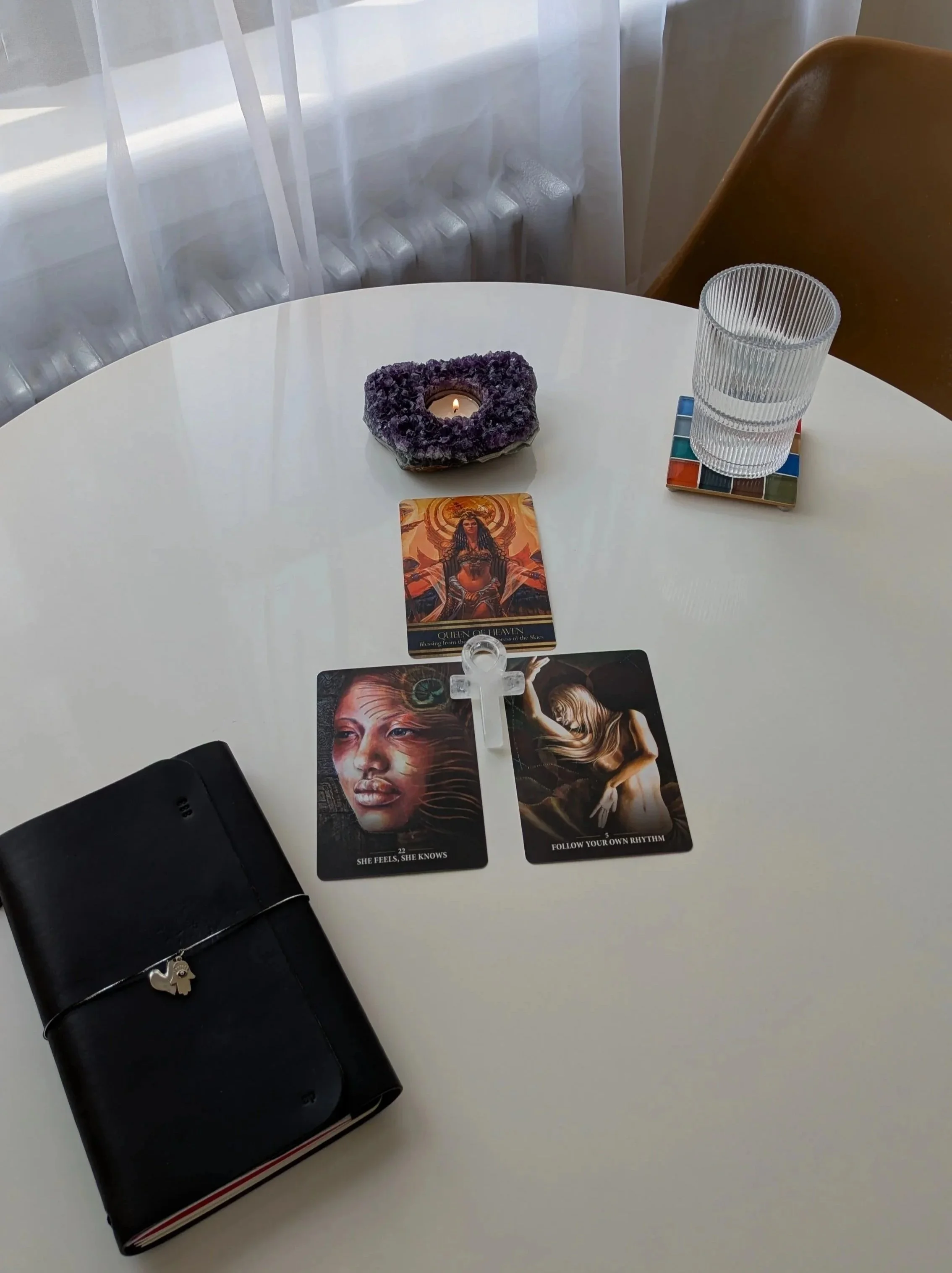A round white table with oracle cards, a candle in amethyst, clear quartz ankh, a glass of water, and a black journal with a hamsa charm. Behind the table is a window with sheer curtains allowing natural light.