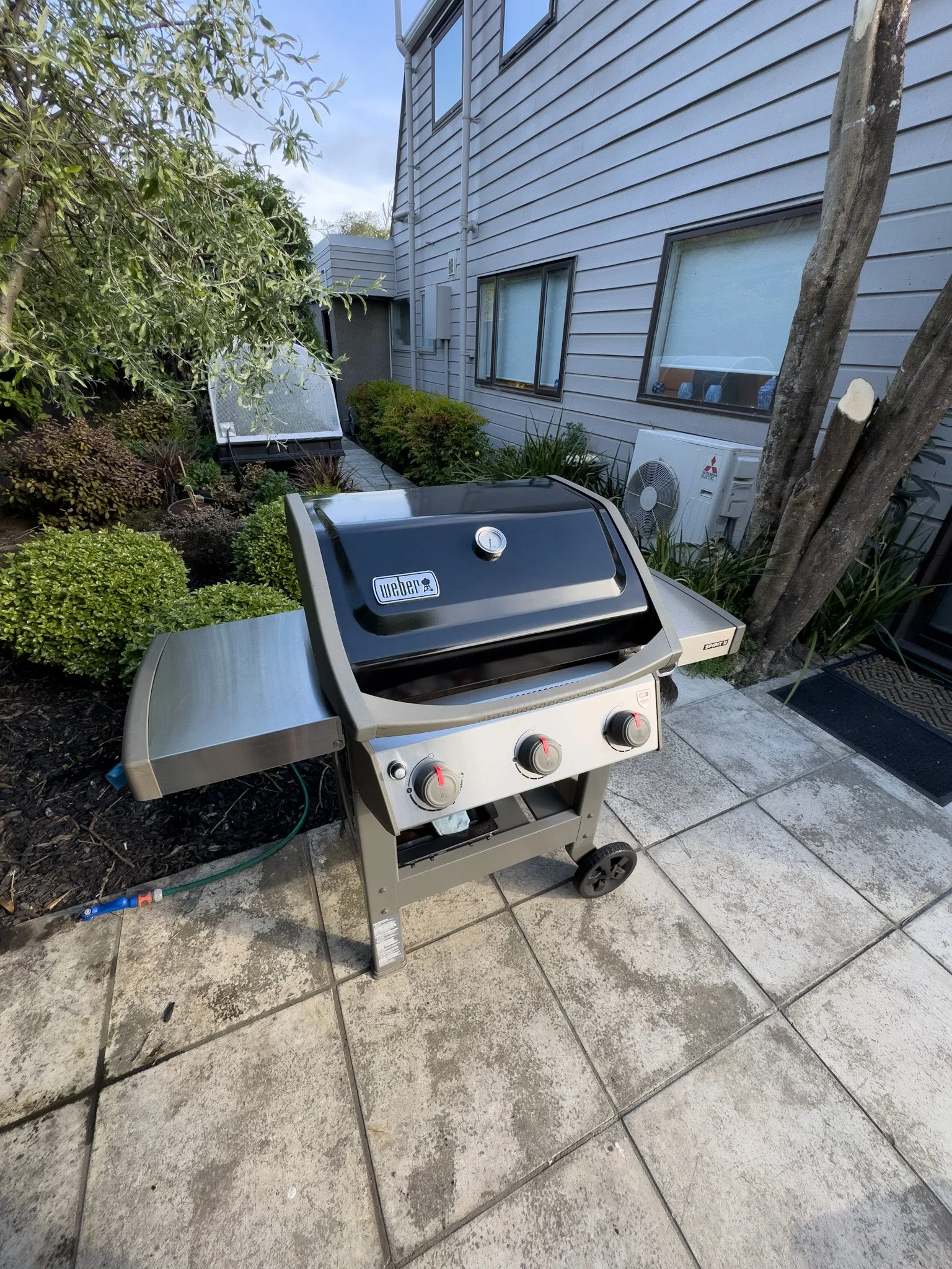 A Weber gas grill on a patio with stone pavers, next to a house with grey siding, a window, and an air conditioning unit, surrounded by plants and bushes.