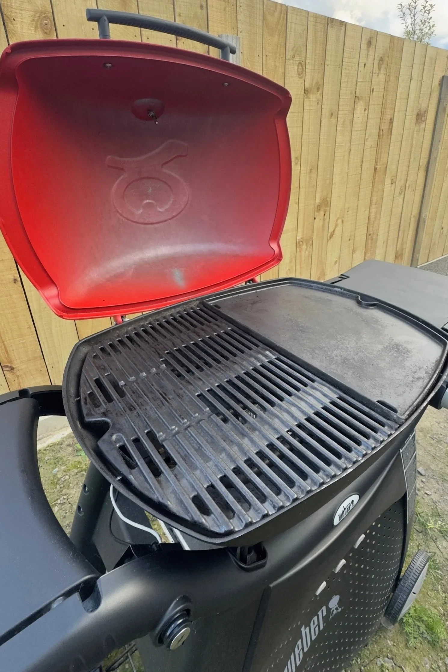 A black Weber grill with a red lid open, showing the grilling surface with some residue, situated outdoors in front of a wooden fence.