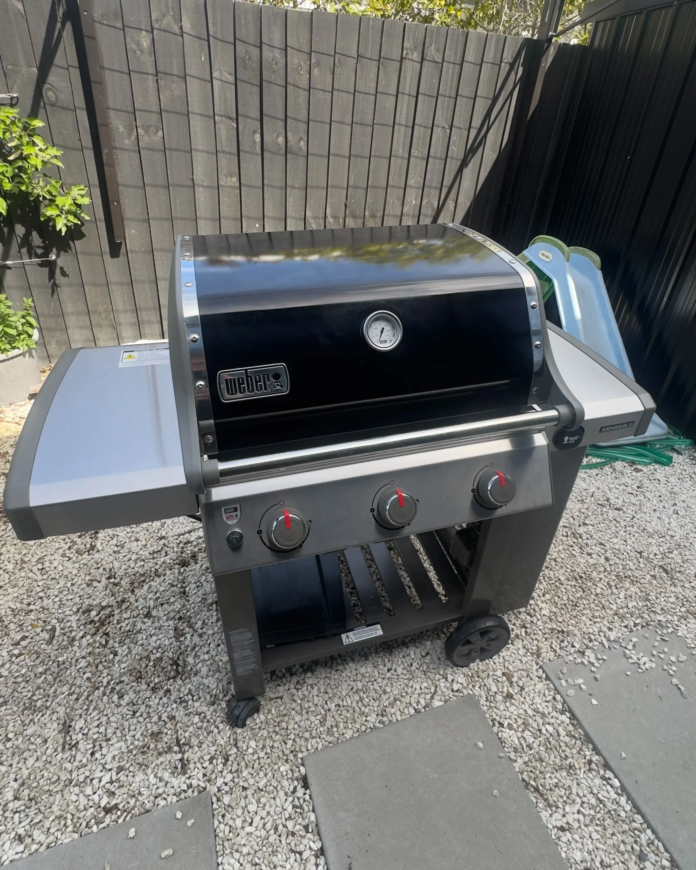 A Weber Genesis gas grill with three control knobs and a thermometer, positioned outdoors on gravel and concrete, with a black fence and blue slide in the background.