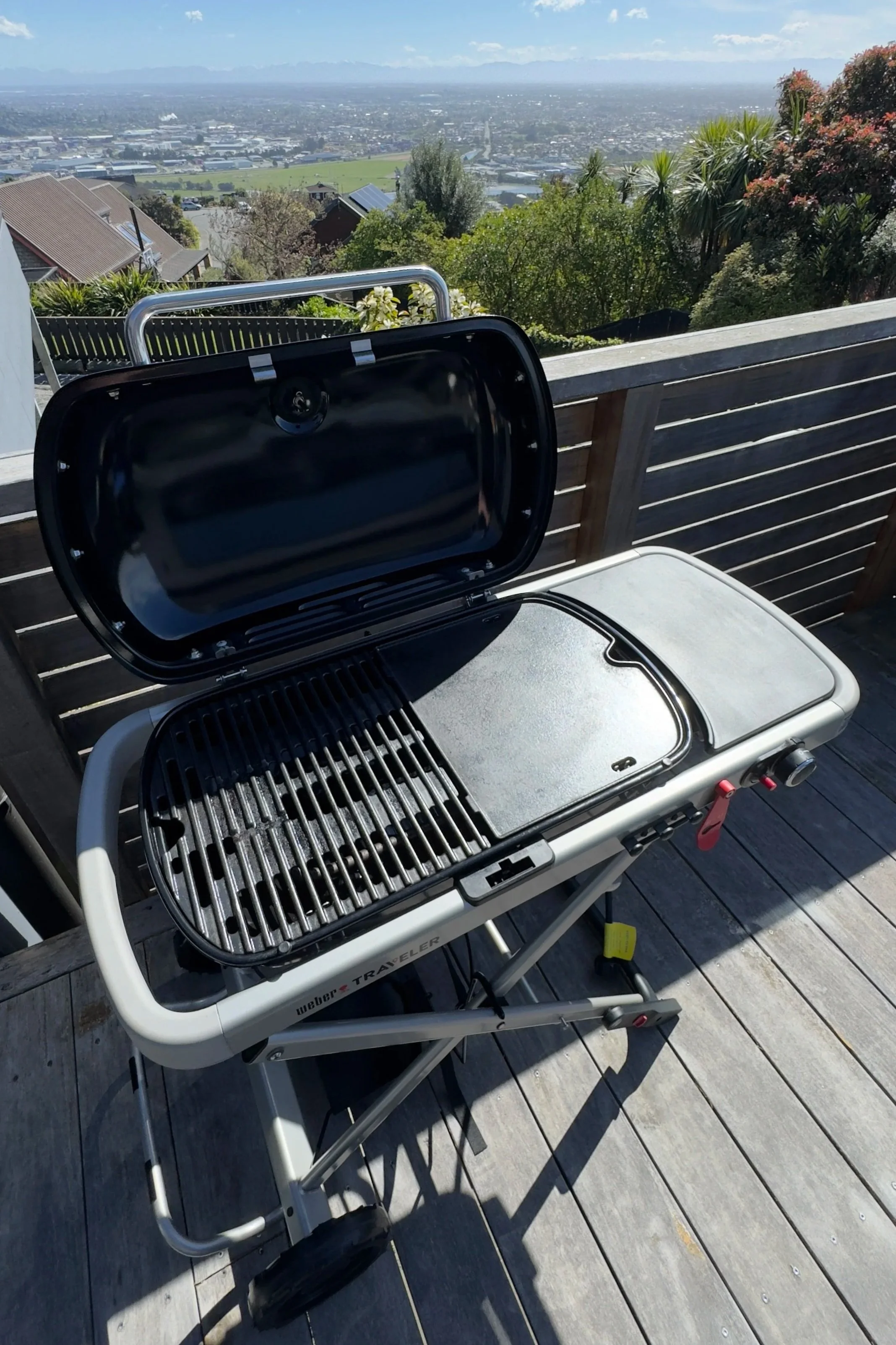 A portable Weber Traveler gas grill with an open lid on a wooden deck, overlooking a cityscape and distant mountains with trees and rooftops in the foreground.