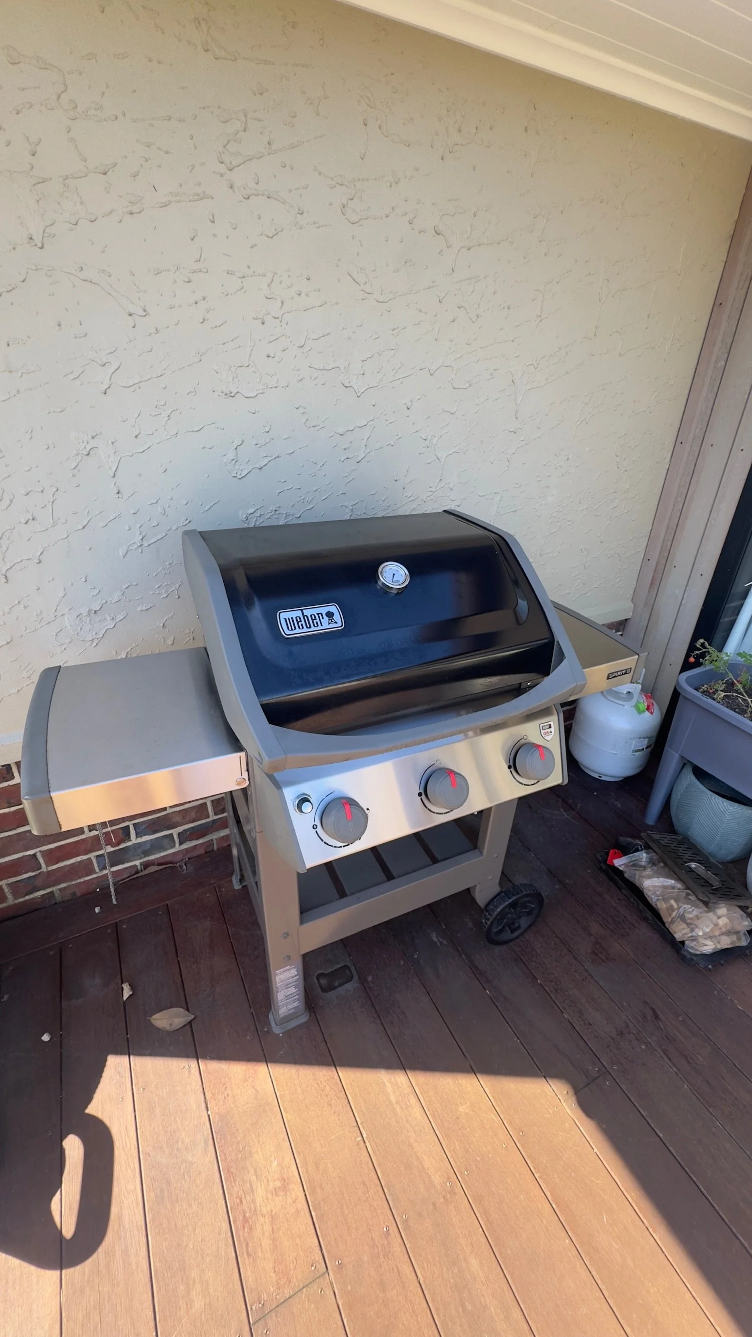 A stainless steel Weber barbecue grill on a wooden patio, with a brick wall and a beige textured wall behind it. There is a propane tank next to the grill on the right side. Part of a gray plant container with plants and gardening tools is visible on the right.