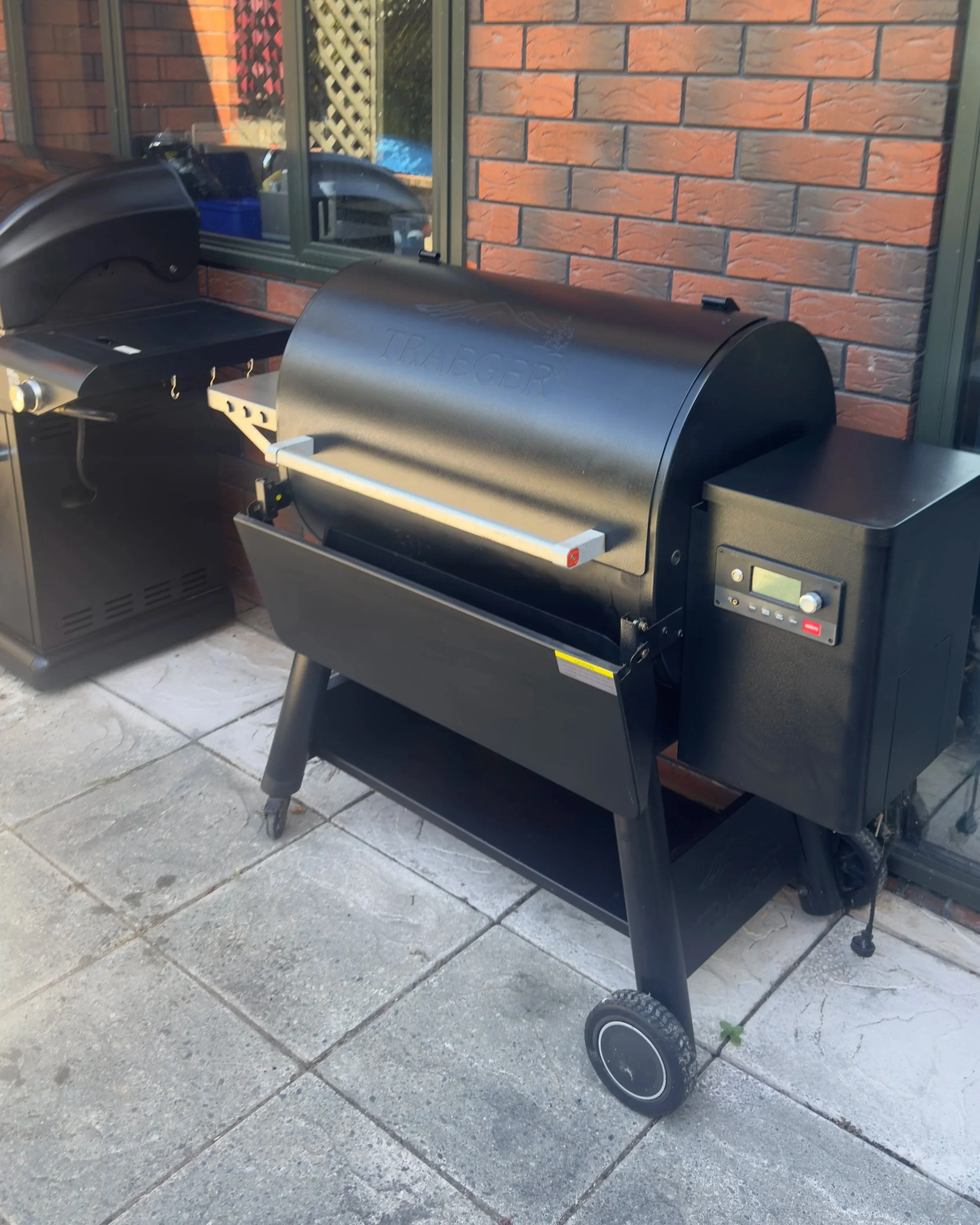Black pellet smoker grill on patio with brick wall background.