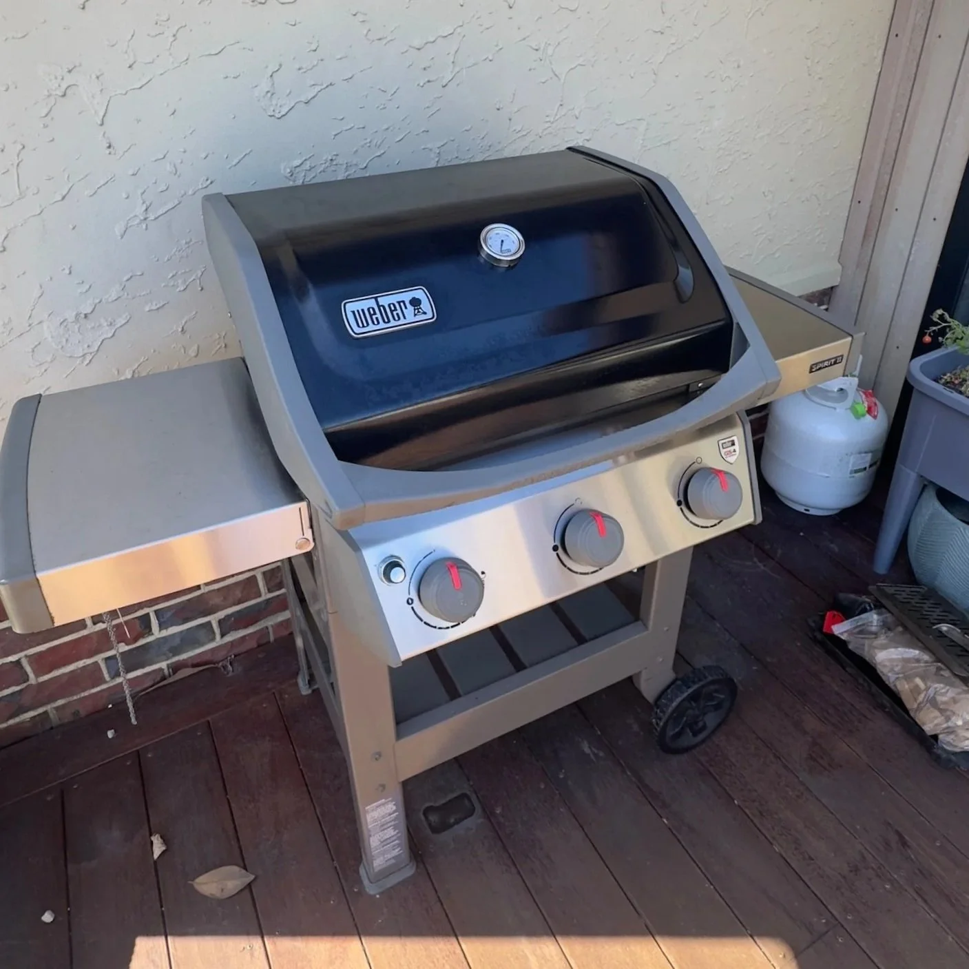 Stainless steel Weber gas grill with black lid and three control knobs, standing on a wooden deck next to a brick wall, with a propane tank nearby.