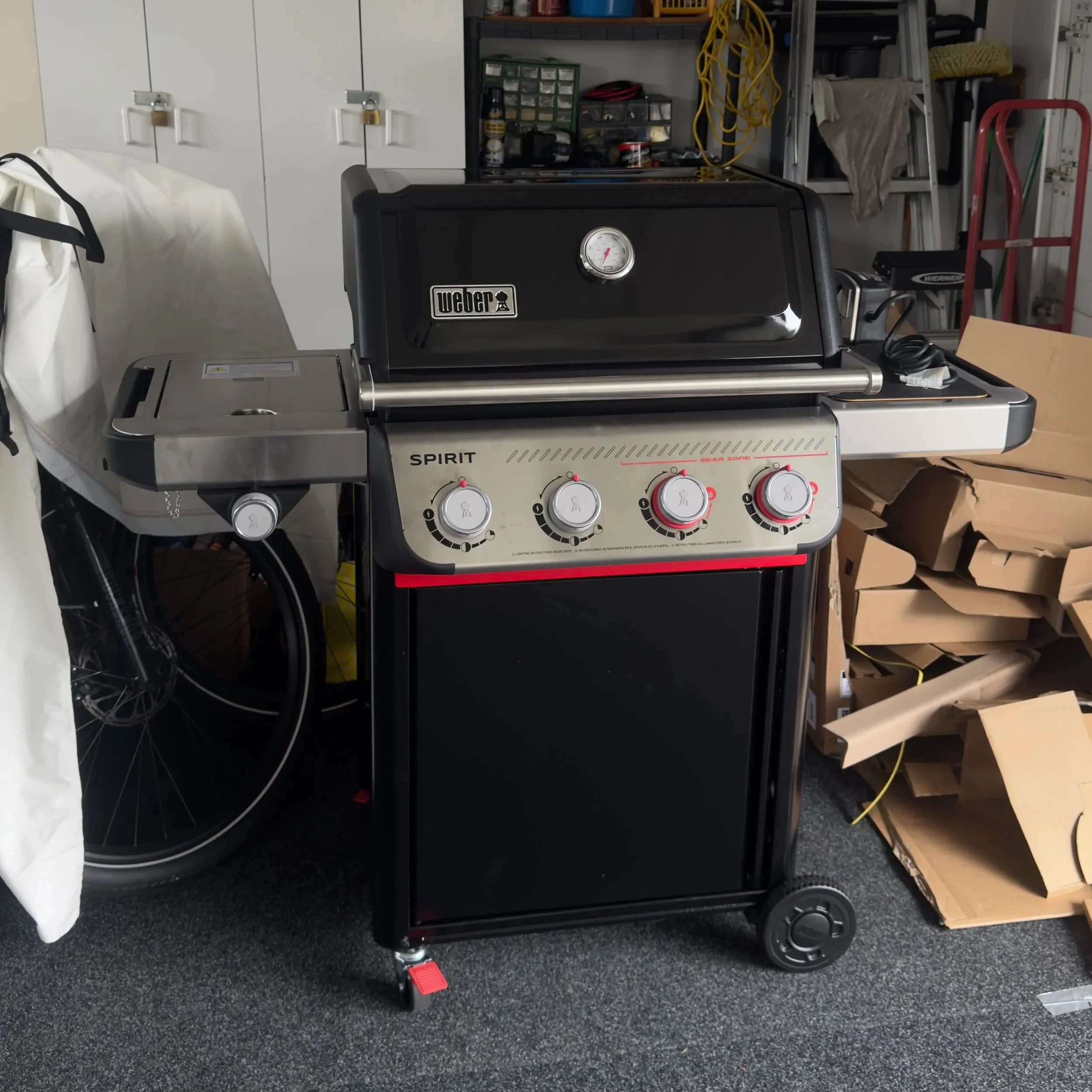 A Weber Spirit gas grill on wheels in a garage with cardboard boxes and shelves in the background.