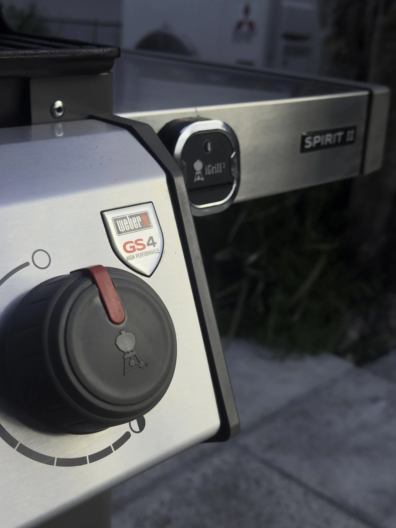 Close-up of a stainless steel grill with a black control knob and a Weber GS4 logo, outdoors with blurred background.