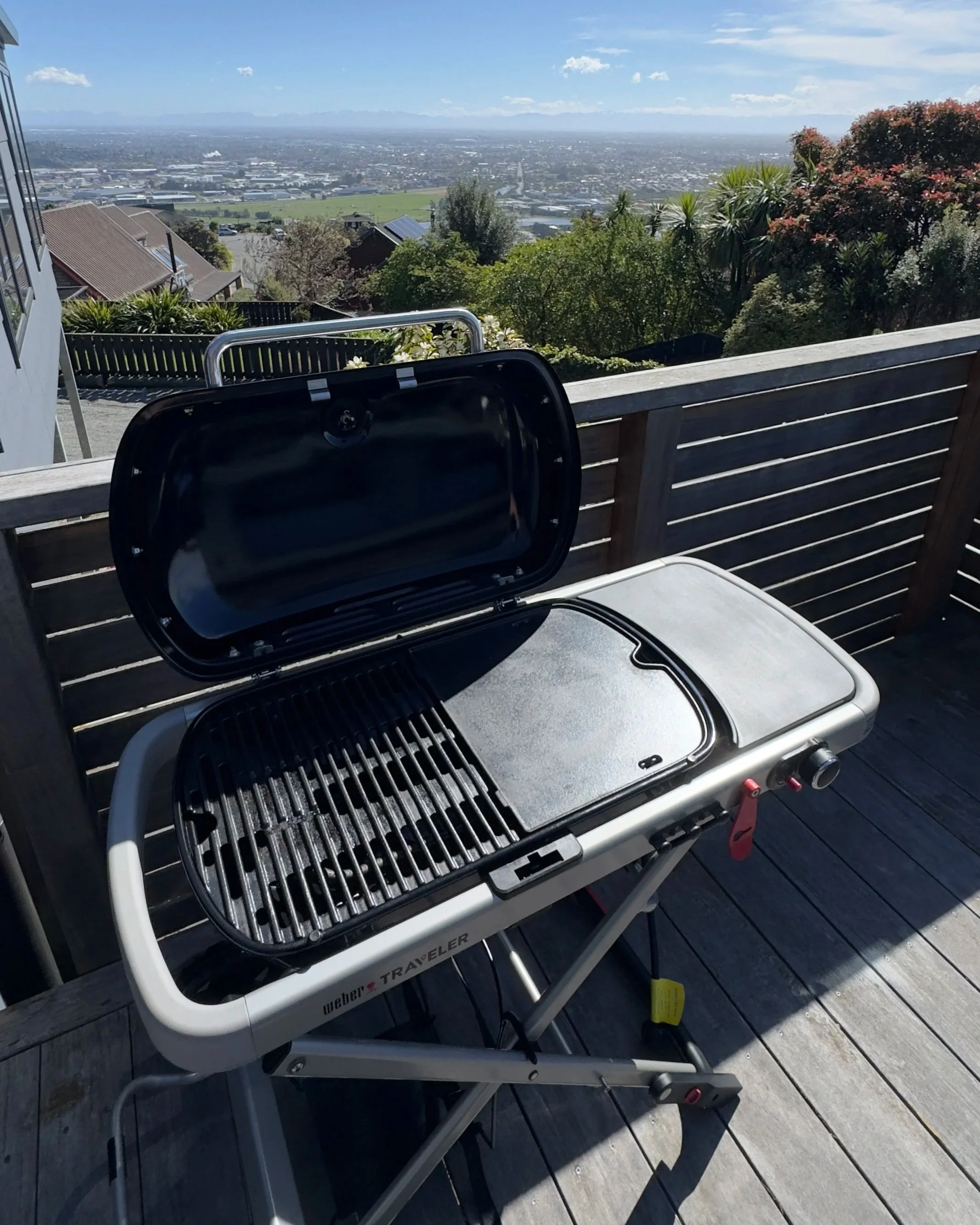 A portable barbecue grill on a wooden deck with a scenic view of a city and mountains in the background. The grill is open, showing the grill grates inside.