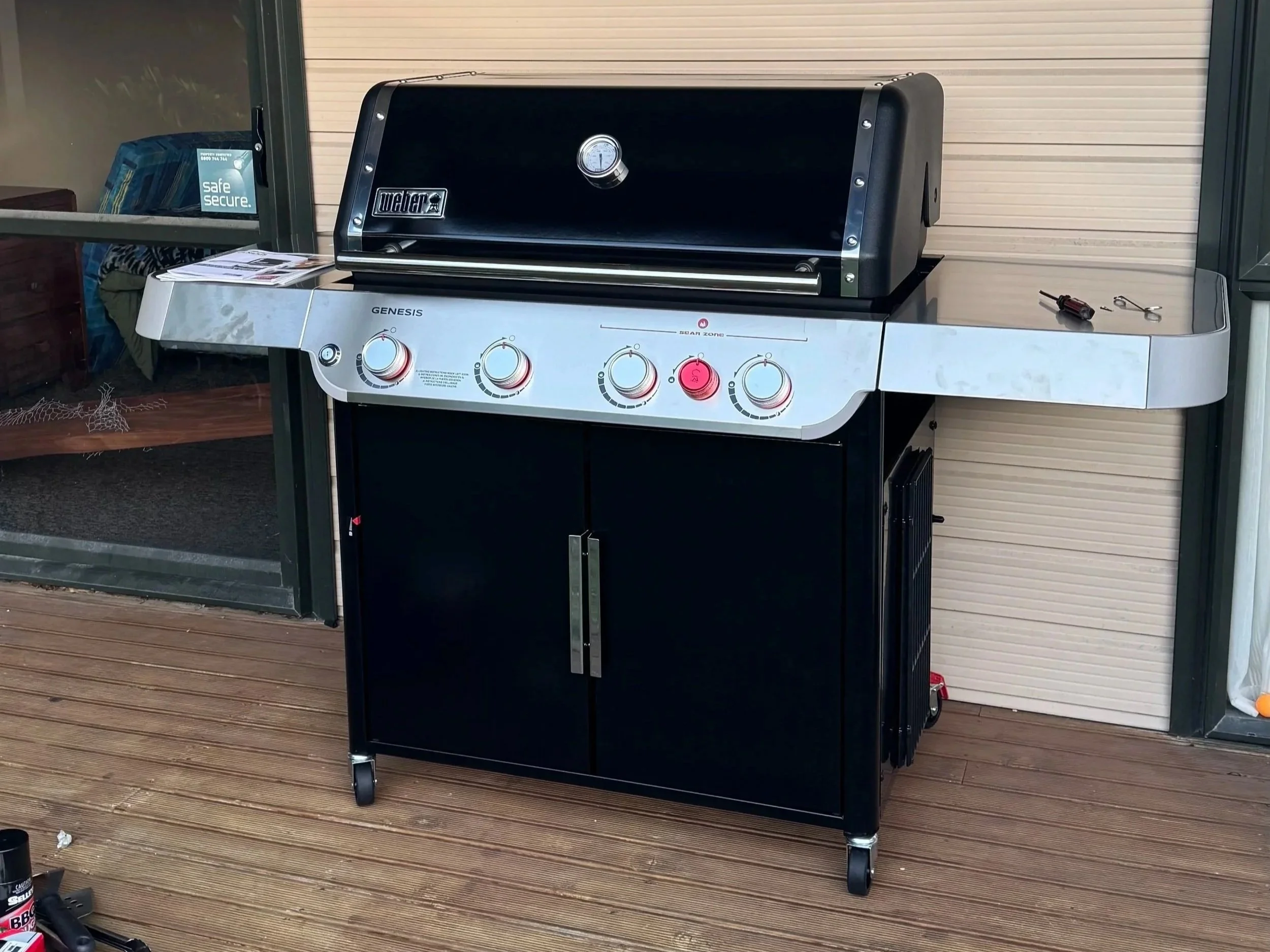 A black outdoor gas grill with a closed lid, four control knobs on the front, and a small thermometer, sitting on a wooden deck.